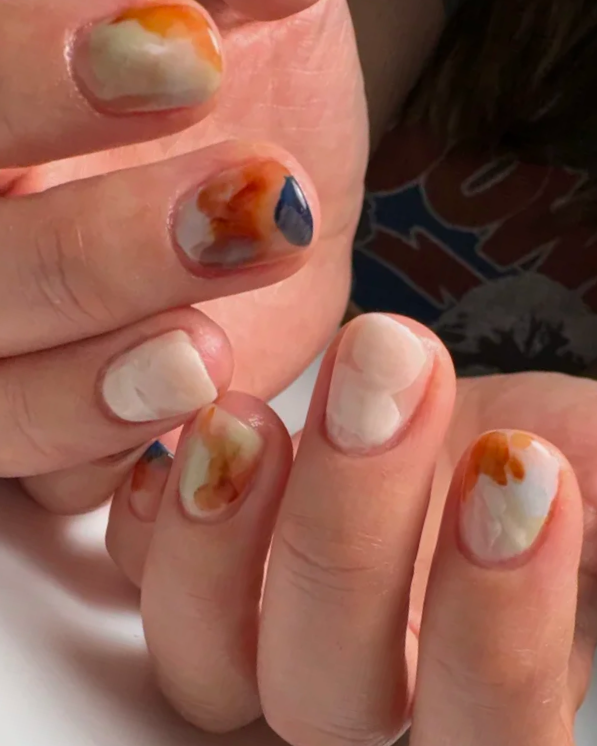 Structured gel manicure featuring abstract nail art in cream, rust, and blue tones.