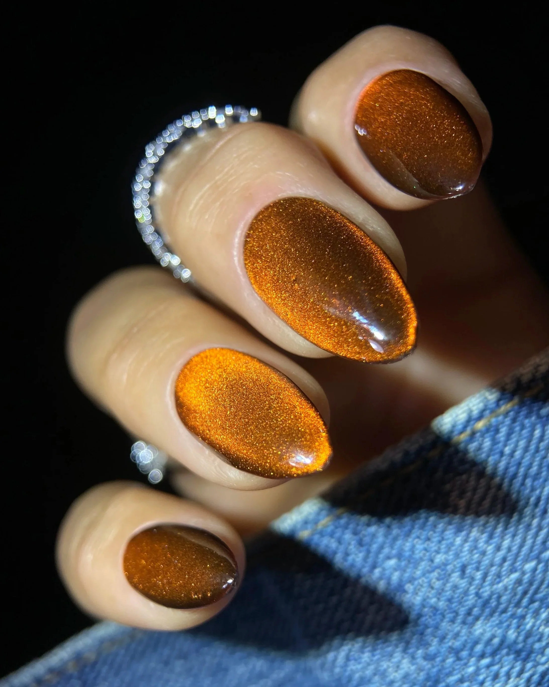 Close-up of Double Rhythm Gemstone Glow MC1329, a warm amber magnet gel with golden honey undertones and a luminous velvet magnetic effect on an almond-shaped nail tip photographed against denim.