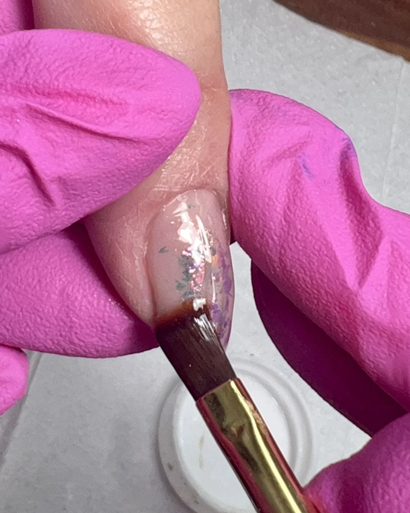 A brush applies clear builder gel over iridescent opal flakes on an almond-shaped nude nail, beginning the encapsulation process before chrome detailing.