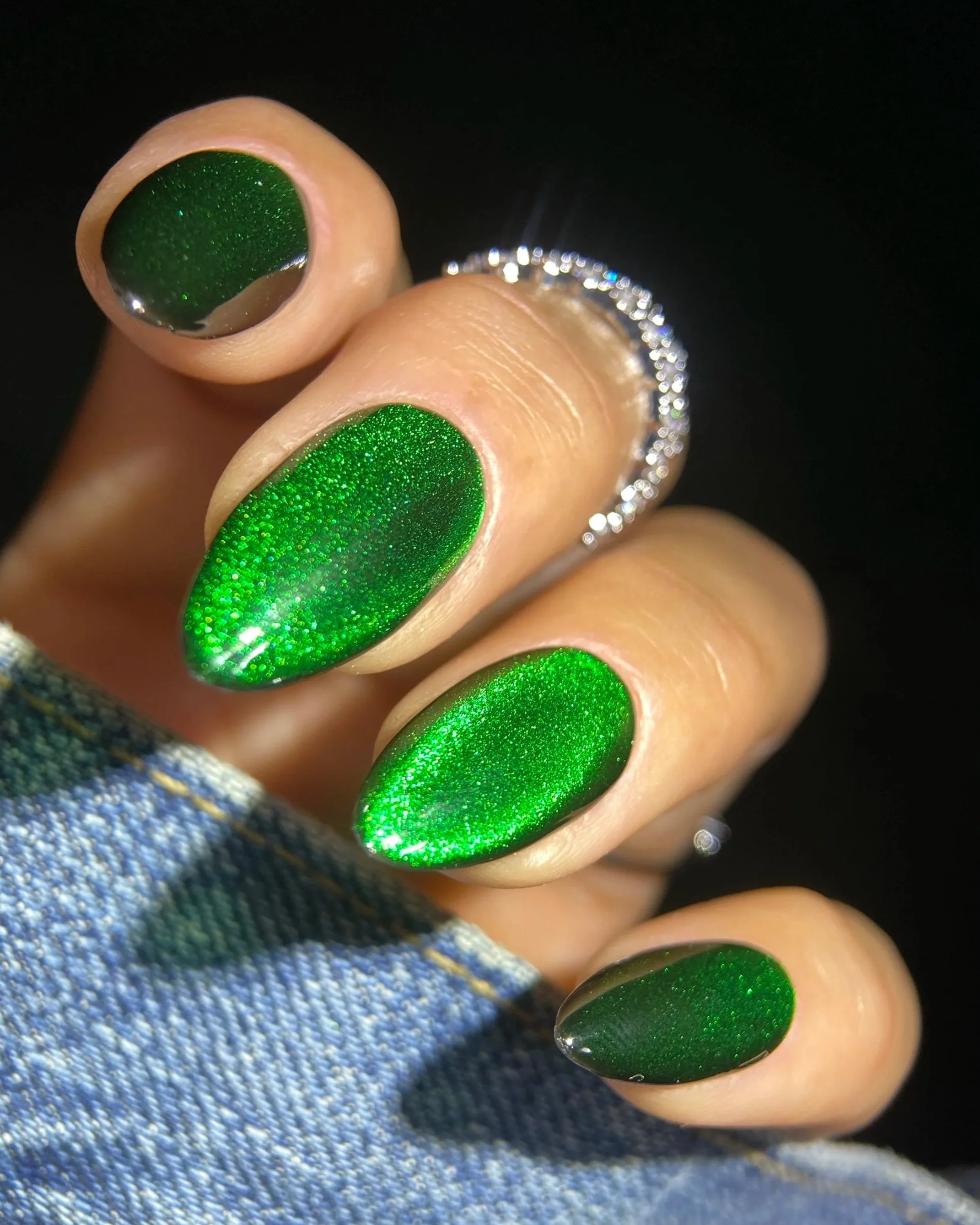 Close-up of Double Rhythm Gemstone Glow MC1326, an emerald green magnet gel with a glowing velvet cat-eye effect and glassy finish on an almond-shaped nail tip against denim.