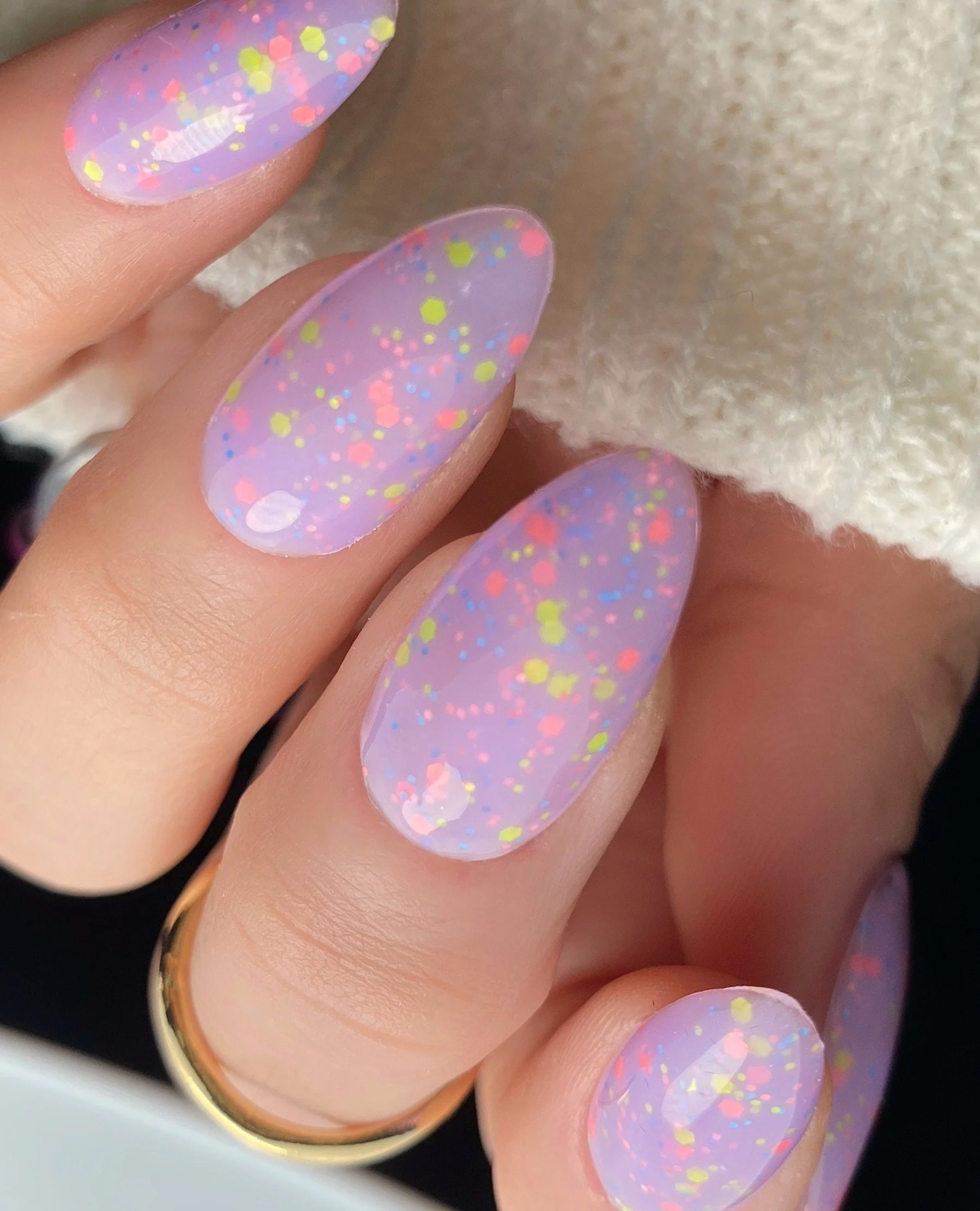 Nail Thoughts Celebration Collection gel color swatch in shade "Extra Sprinkles" a milky lavender with colorful satin glitter on manicured hand with rings and knit sleeve.