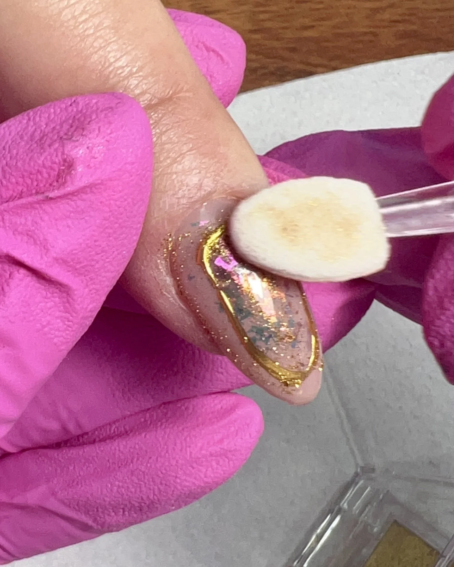 A sponge applicator buffs brassy gold chrome powder onto a cured oval outline surrounding encapsulated opal flakes on a nude almond-shaped nail.
