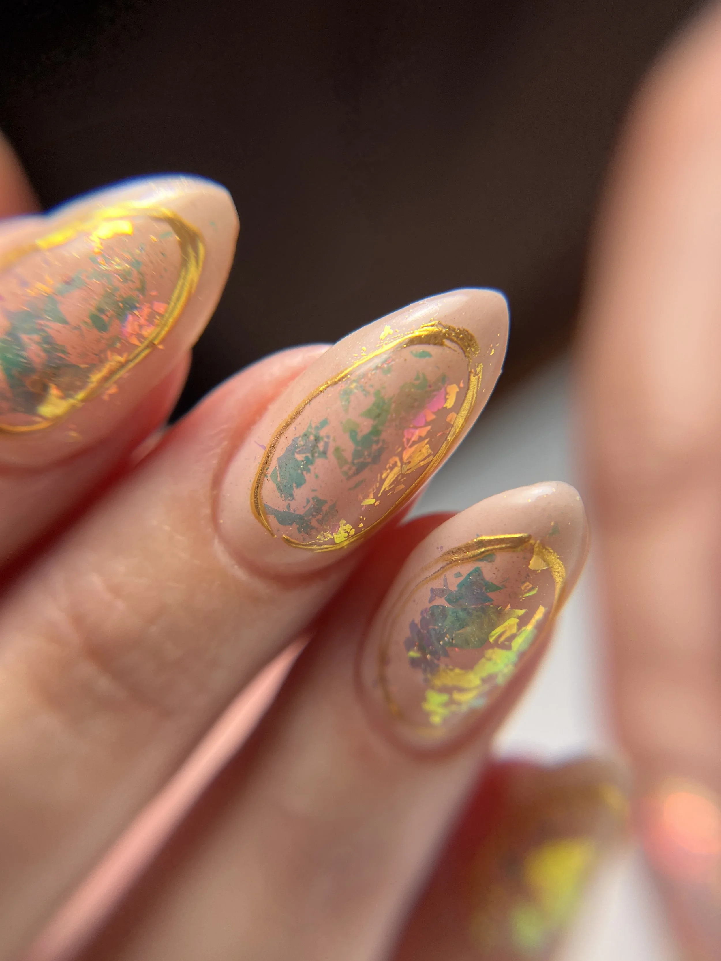 Close-up of almond-shaped nude nails featuring encapsulated iridescent opal flakes framed with thin brassy gold chrome ovals. The flakes shift pink, gold, green, and blue beneath a glossy gel finish.