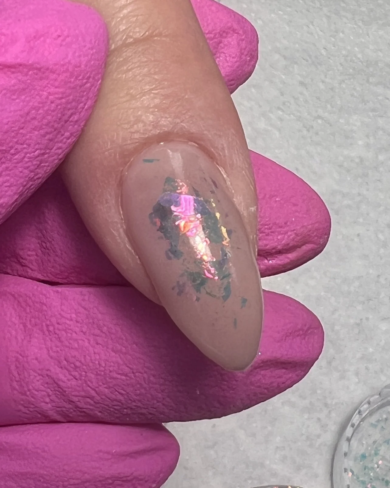 Close-up of an almond-shaped nude nail with iridescent opal flakes pressed into the sticky layer of gel. The flakes reflect pink, gold, and green tones against a sheer neutral background.