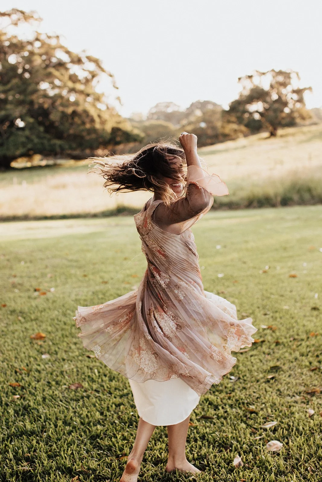 A woman with curly hair spinning in a grassy field, wearing a flowy floral dress, with trees in the background and sunlight.
