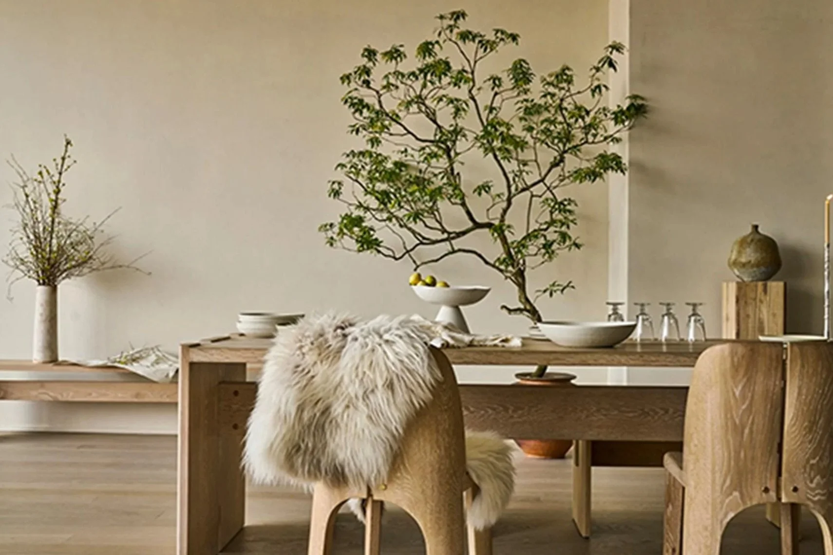 Wood dining table with a fur throw draped over the back of a wooden chair, decorated with plants and bowls, against a plain wall.