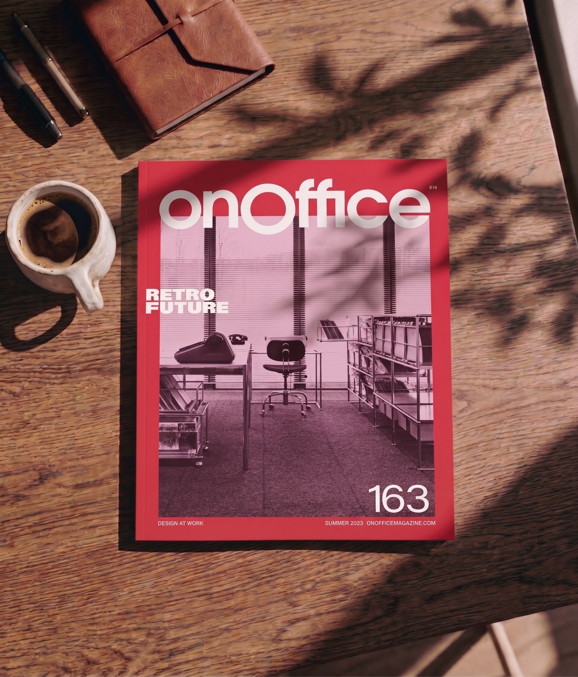 Close-up of a wooden table with a magazine titled 'OnOffice' featuring office design images, a ceramic mug with coffee, two pens, and a leather-bound notebook.