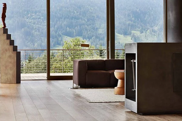 Modern living room with large glass windows overlooking a mountain view, brown sofa, wooden side table, and a floor lamp.