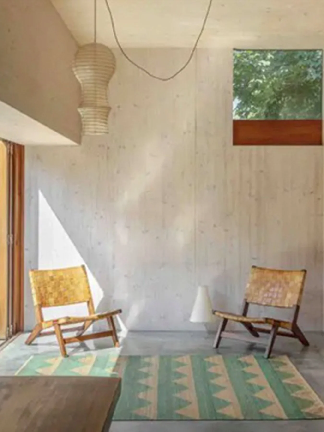 A room with two wooden chairs with woven seats, a white cone-shaped lamp, a window overlooking green trees, a colorful patterned rug, and a light-colored wall and ceiling.