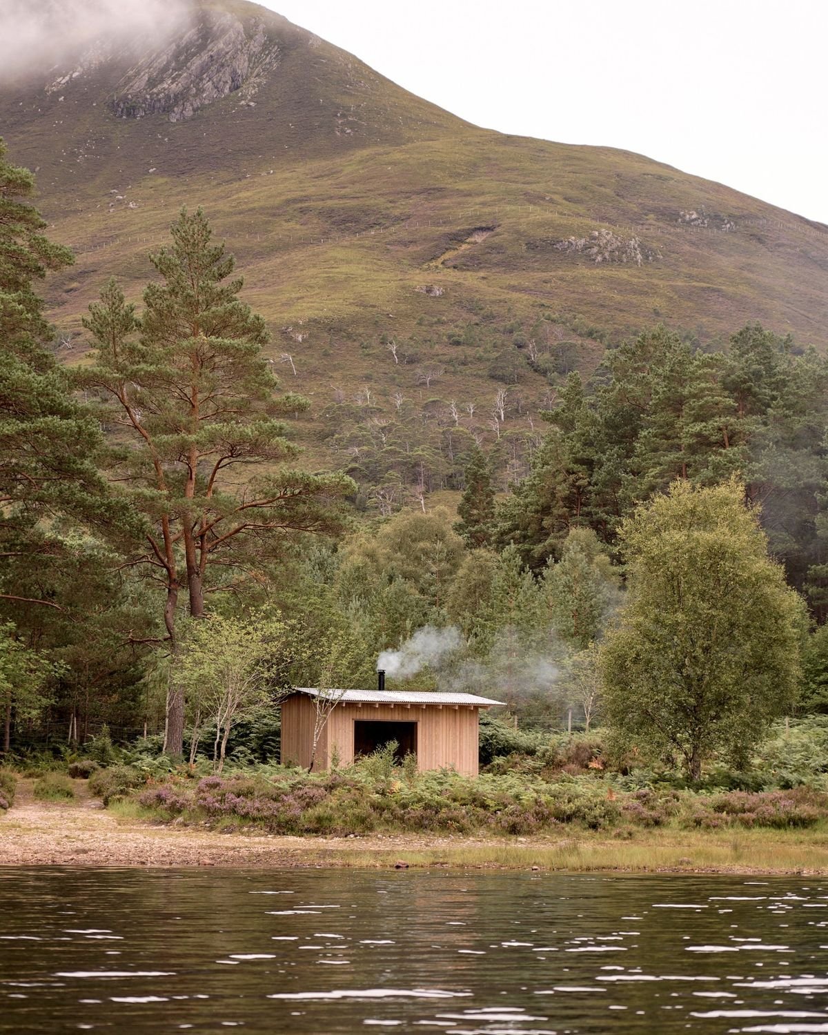 A small wooden cabin with a chimney emitting smoke, surrounded by trees and near a body of water, with a large mountain in the background.