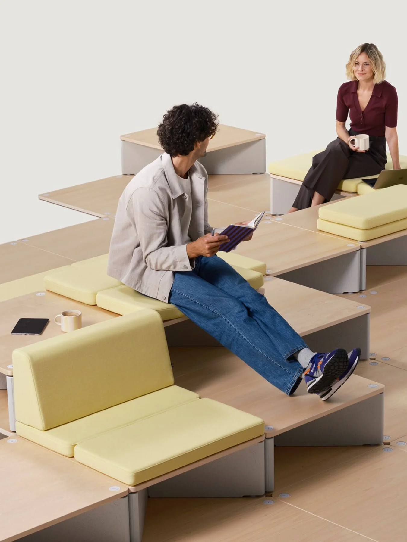 A man with curly hair sitting on a yellow cushioned bench, reading a notebook, in a modern office space. A woman with blonde hair, wearing a burgundy top and black pants, is sitting on another bench holding a mug, with a laptop nearby. The room has light wood tables with yellow cushions and a neutral background.