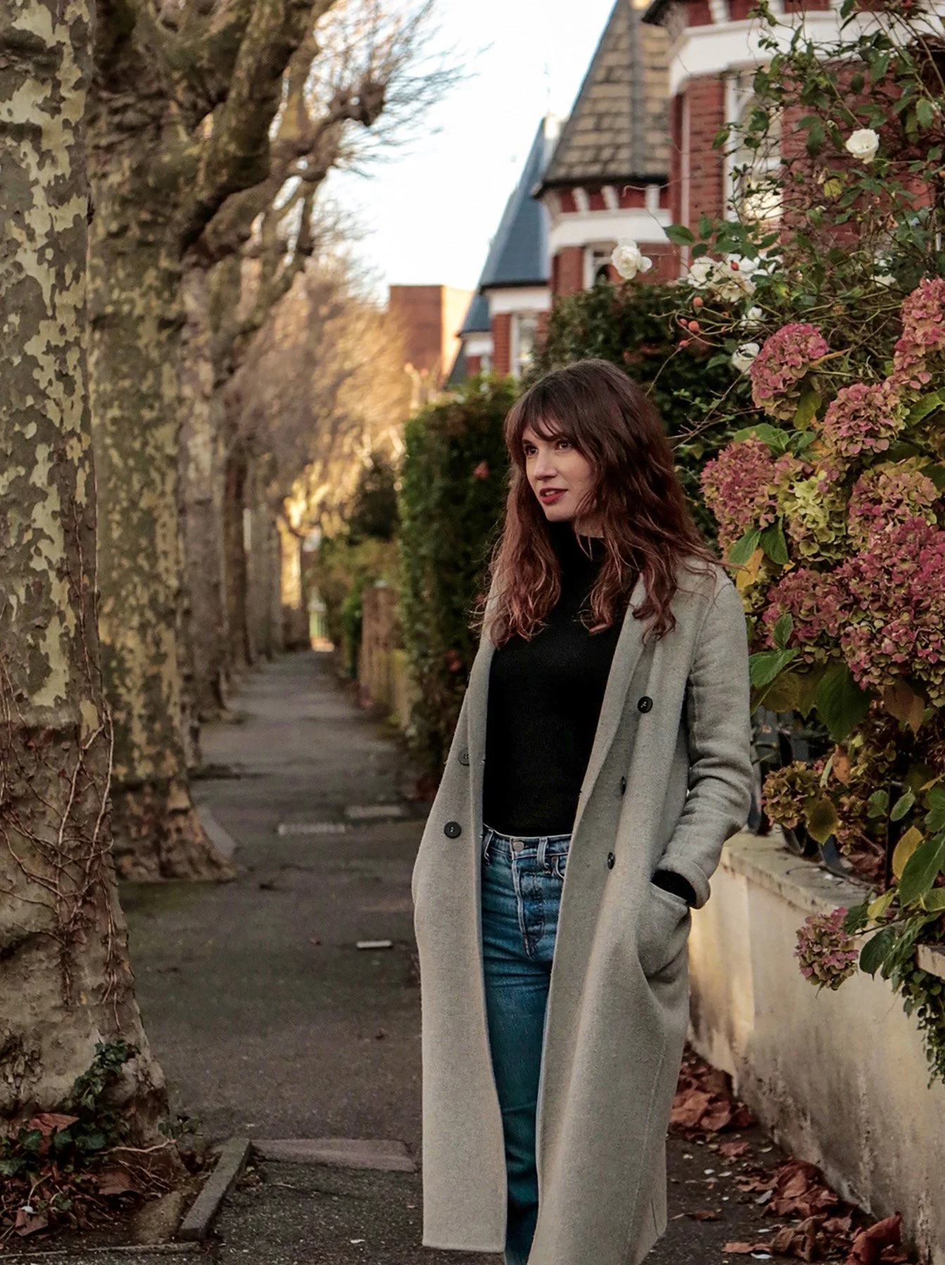 A woman with wavy brown hair and red lipstick standing on a sidewalk surrounded by trees and flowering bushes, wearing a long gray coat, black top, and blue jeans. Houses with Victorian architecture are visible in the background.