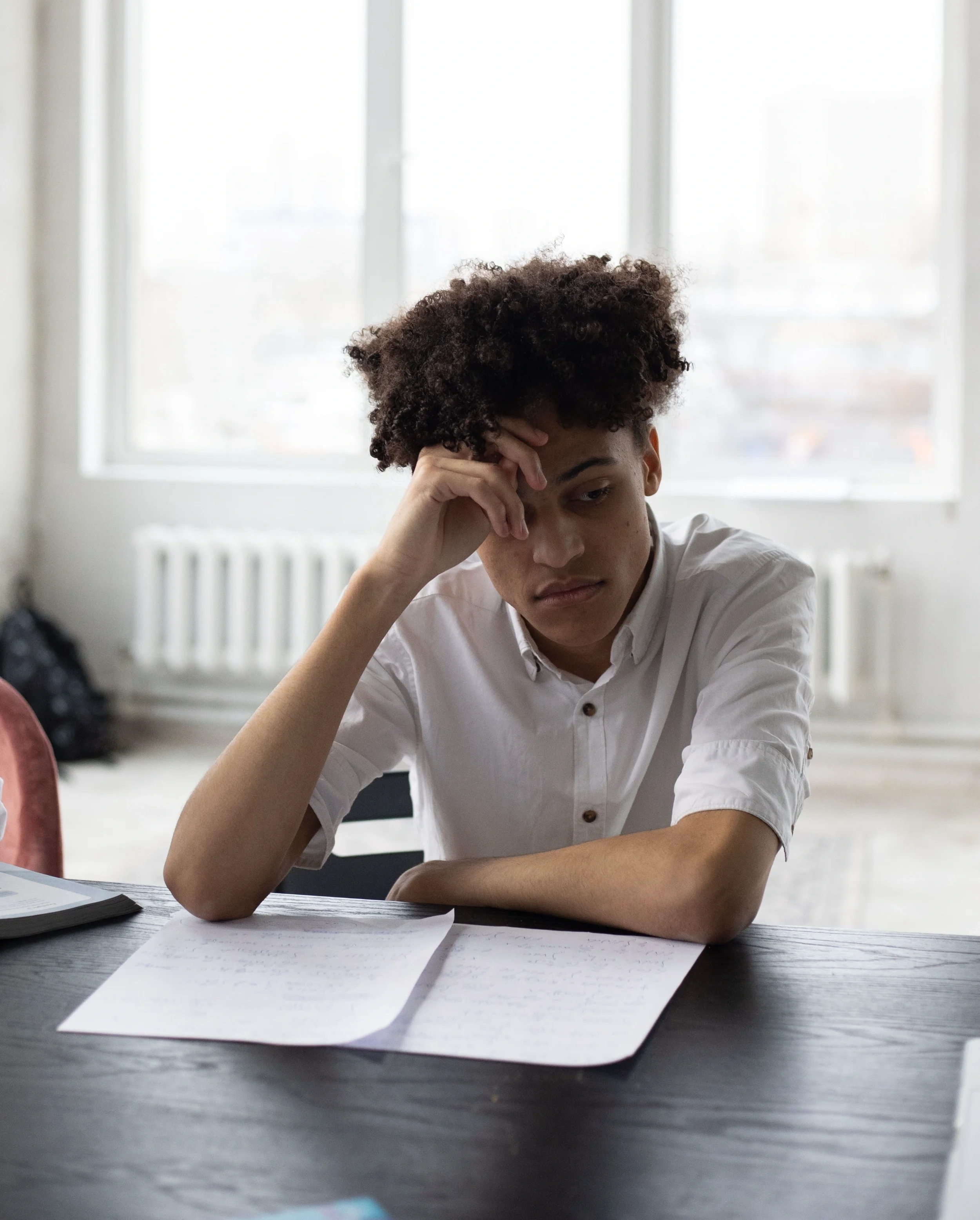 A young man with curly dark hair sitting at a desk, resting his head on his hand.