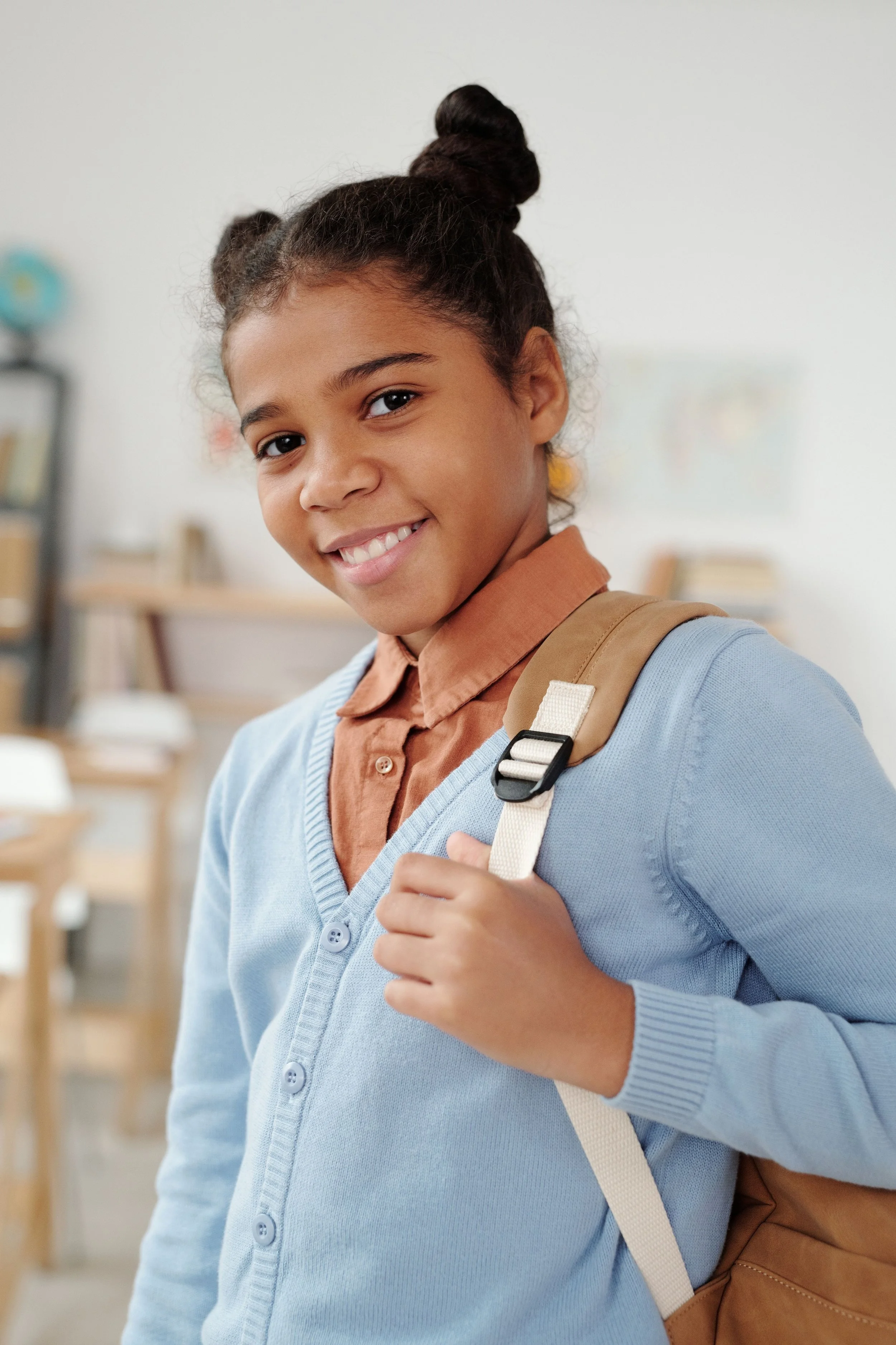 A young girl with dark hair smiling, and carrying a tan backpack.
