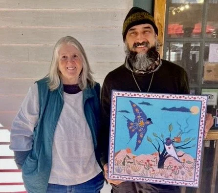 Two people, a woman and a man, smiling while standing outdoors. The man is holding a colorful painting of a tree, and two birds. The woman is wearing a light-colored shirt and blue vest, and the man is wearing a black hoodie and beanie.