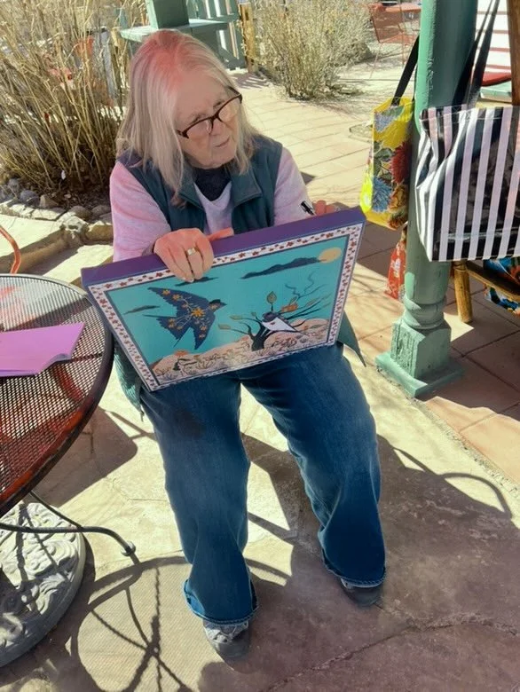 Kate Lindsey sitting outdoors, signing the back of a colorful canvas with a bird on the front.