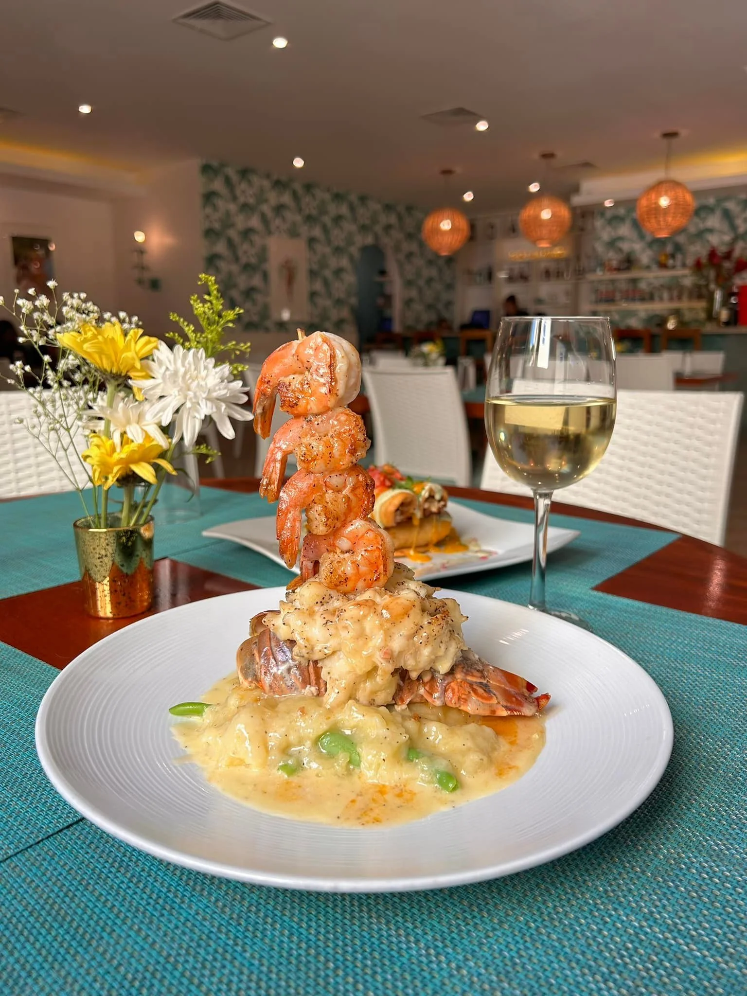 A gourmet seafood dish featuring a tall skewer of grilled shrimp over lobster tail and risotto, served with a glass of white wine at Red Ginger restaurant in San Pedro, Belize.