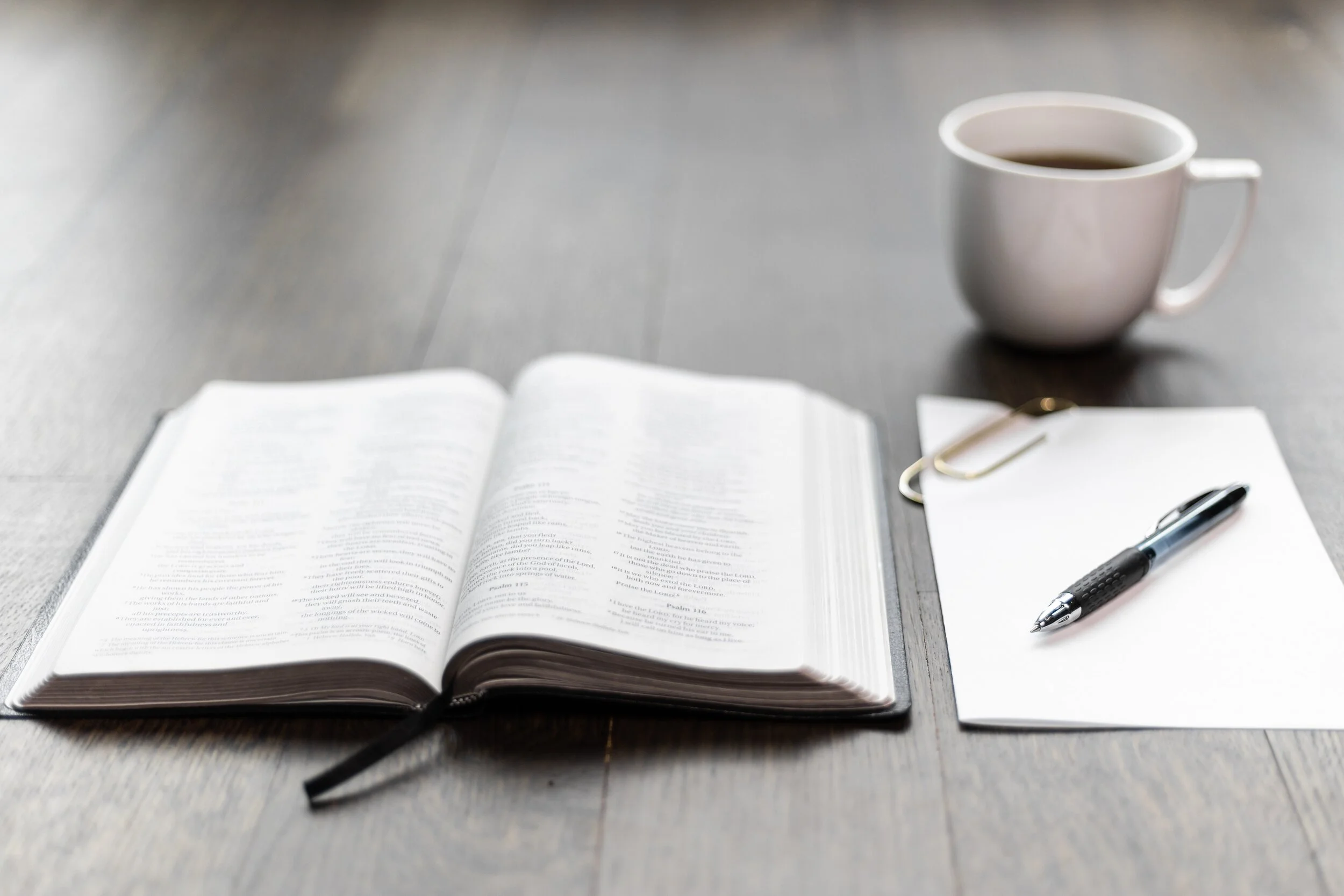 Open Bible on a wooden table with a black and silver pen, a paperclip, a notebook, and a white coffee mug filled with black coffee.
