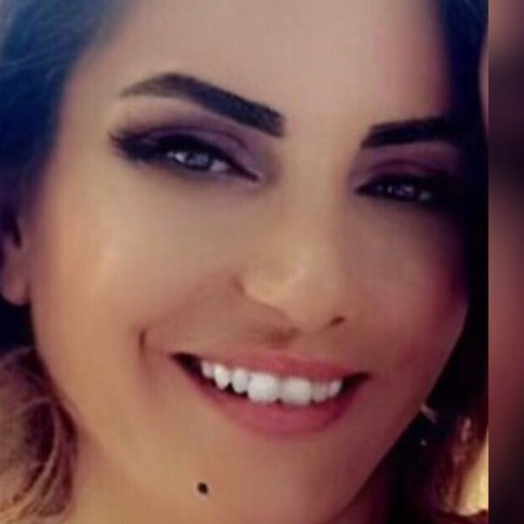 Close-up of a smiling woman with dark hair, dramatic eye makeup, and a beauty mark below her lower lip.