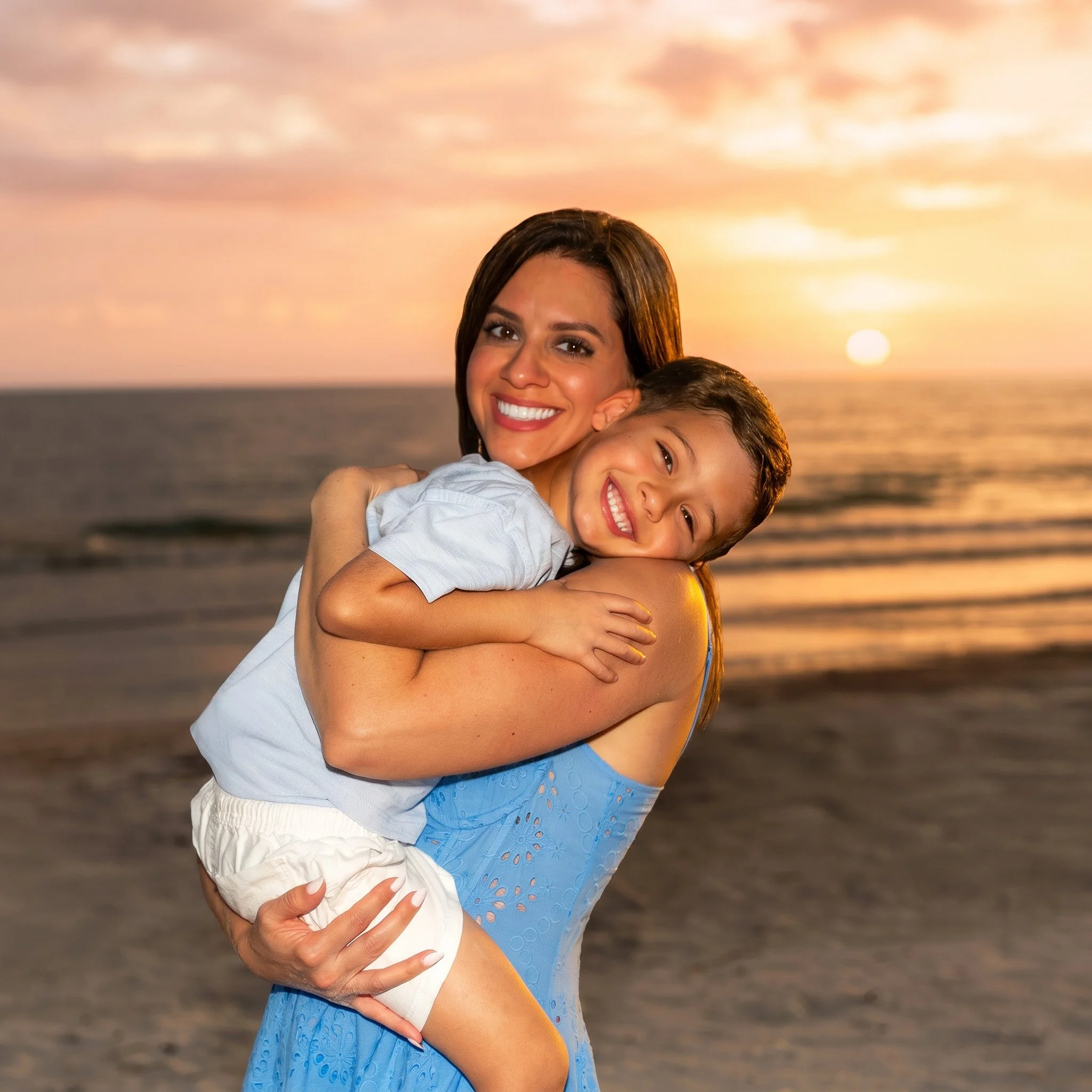 A woman and a young girl smiling and hugging at the beach during sunset.