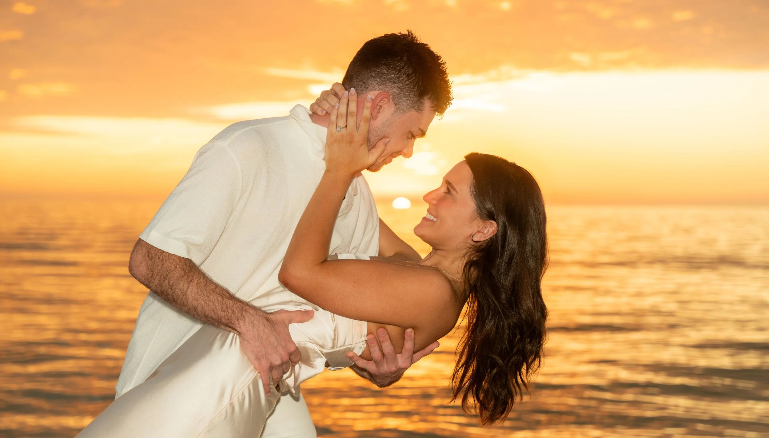 A couple dancing by the water at sunset, with the man holding the woman and both smiling at each other.