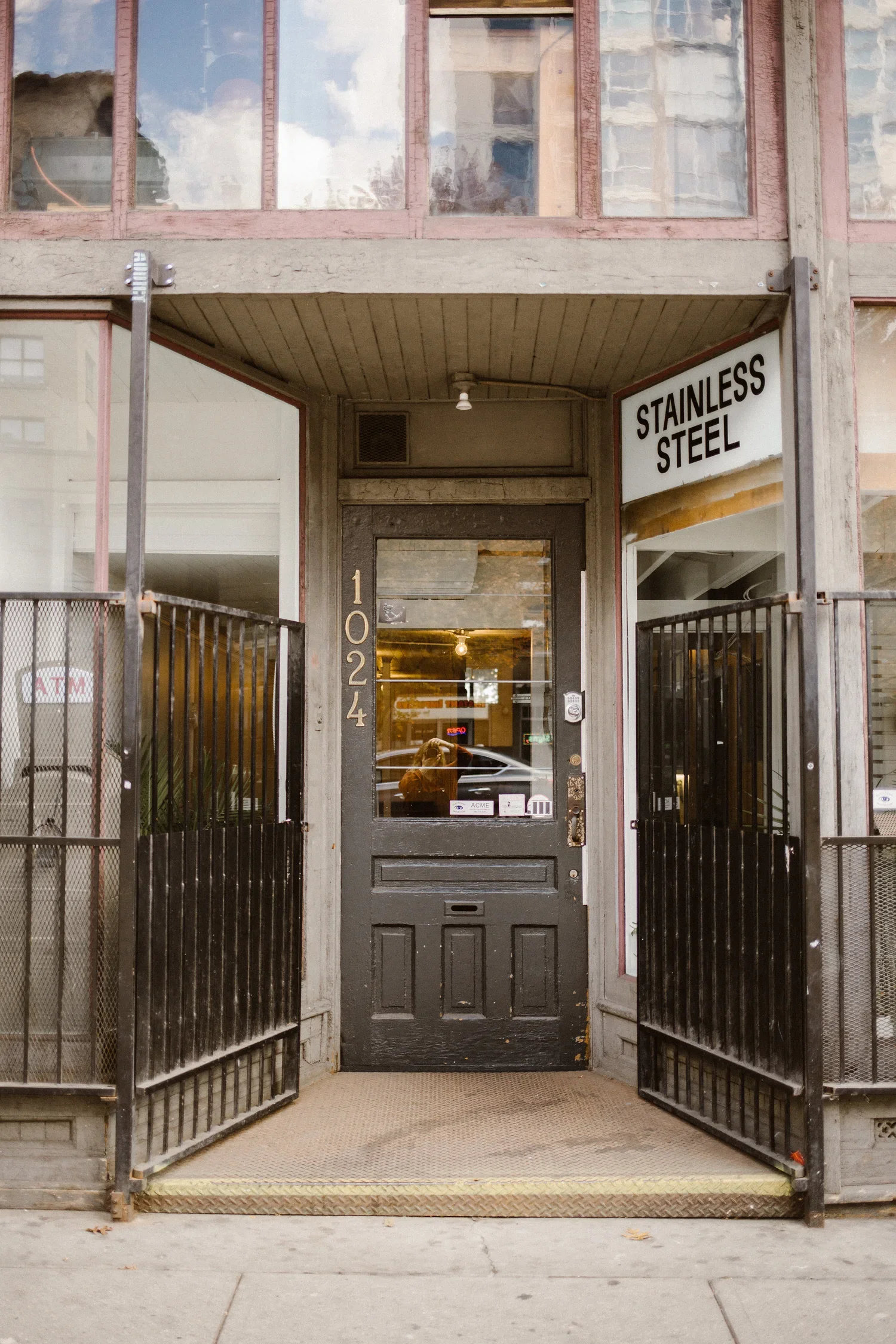 Entrance to a building with a black door, the address '1024' on the left side, a sign that reads 'Stainless Steel,' and open black gates on either side.