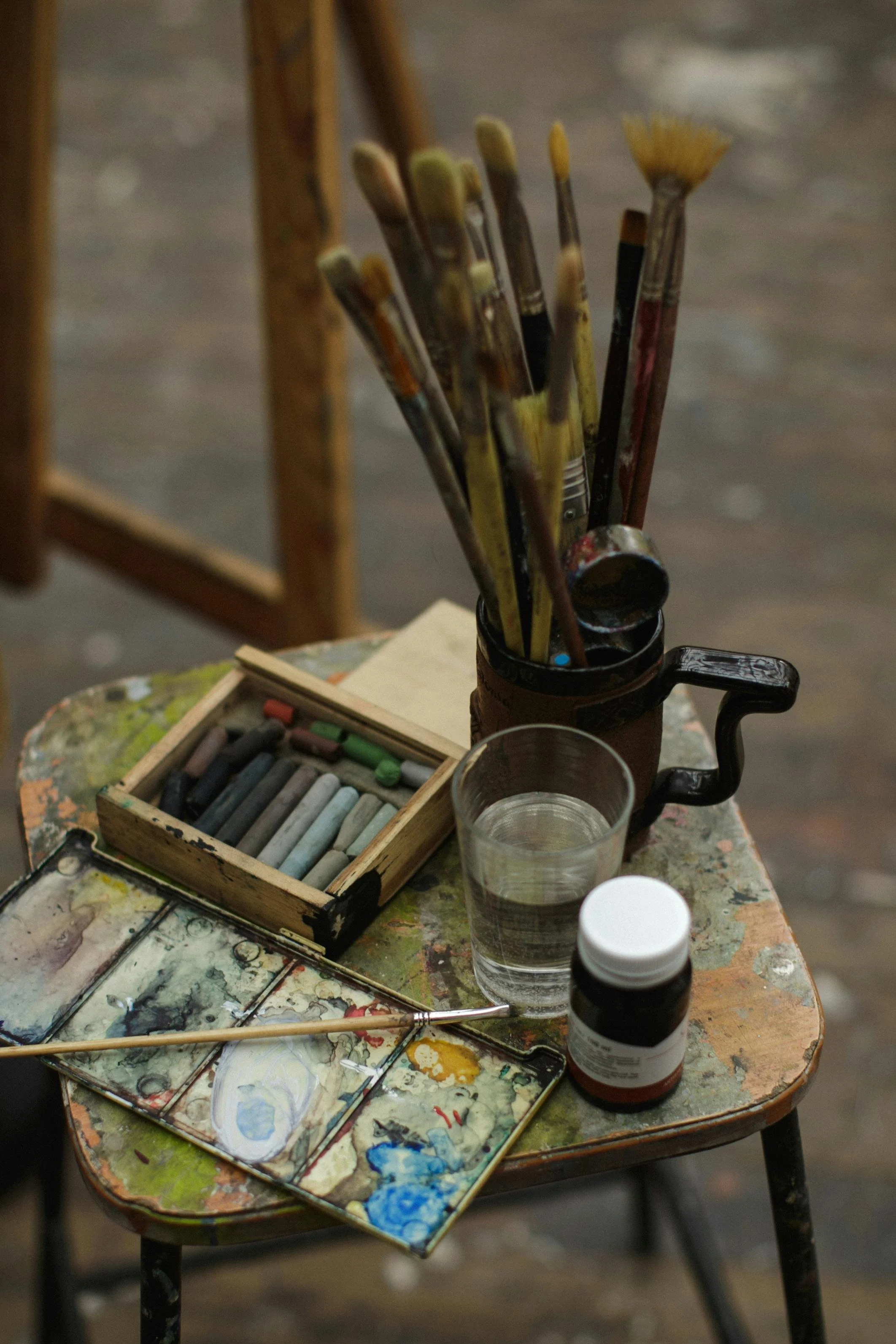 Art supplies on a wooden chair, including a palette with paint, a jar of water, a set of pastels, paintbrushes, and a bottle of paint.