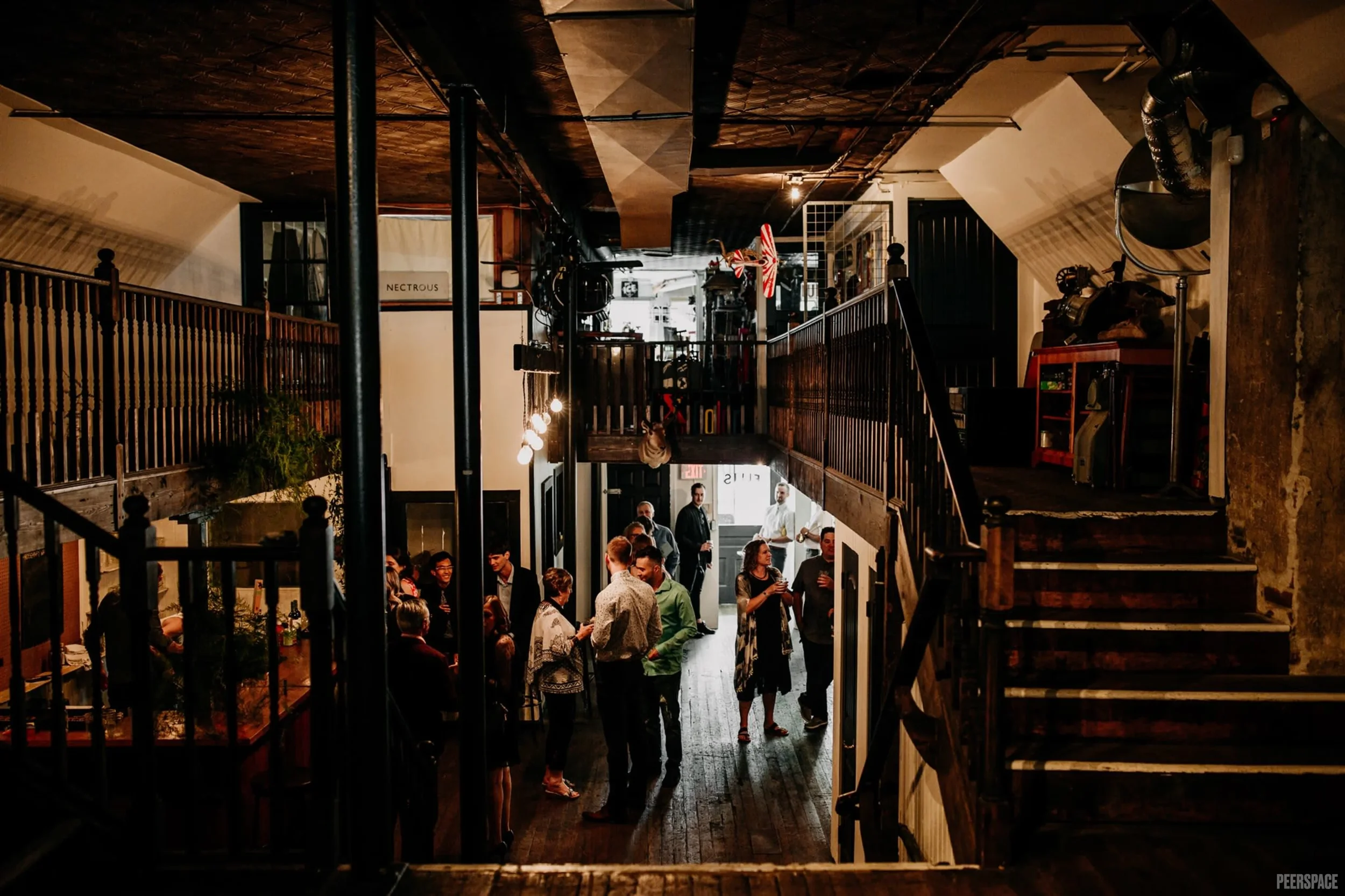 People socializing inside a rustic, multi-level bar or restaurant with wooden stairs and railings, exposed brick and industrial-style lighting. Vancouver event space for rent.