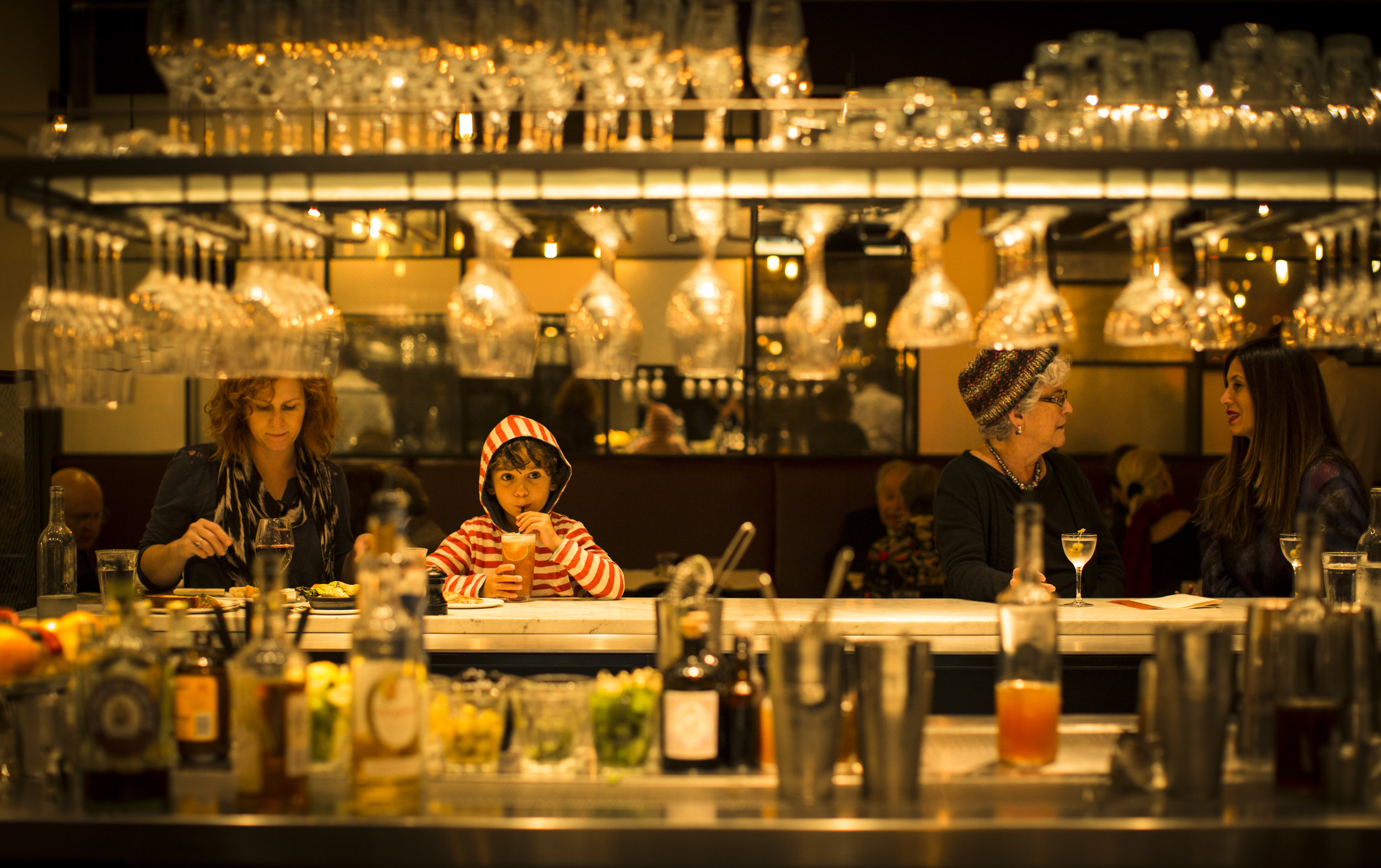  Woman and child sipping juice sit at left side of bar, two women chatting sit on the right side, hanging glasses, bottles, and bar equipment in foreground 