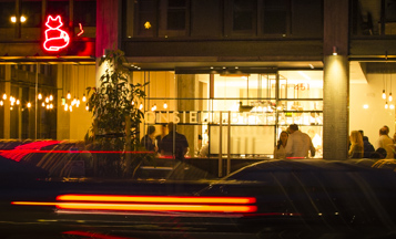  restaurant exterior with glass windows, warmly lit from within, a red neon silhouette of fox above, blurred lights from moving cars streak across 