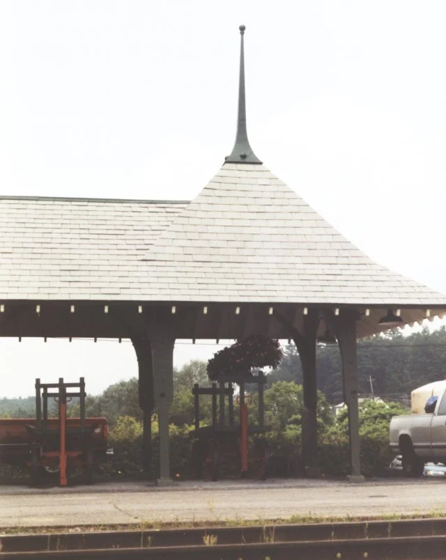 Roof details at the north end of the passenger shelter. A baggage wagon is parked in under. The indistinct blobs are large flower planters hanging from the roof.