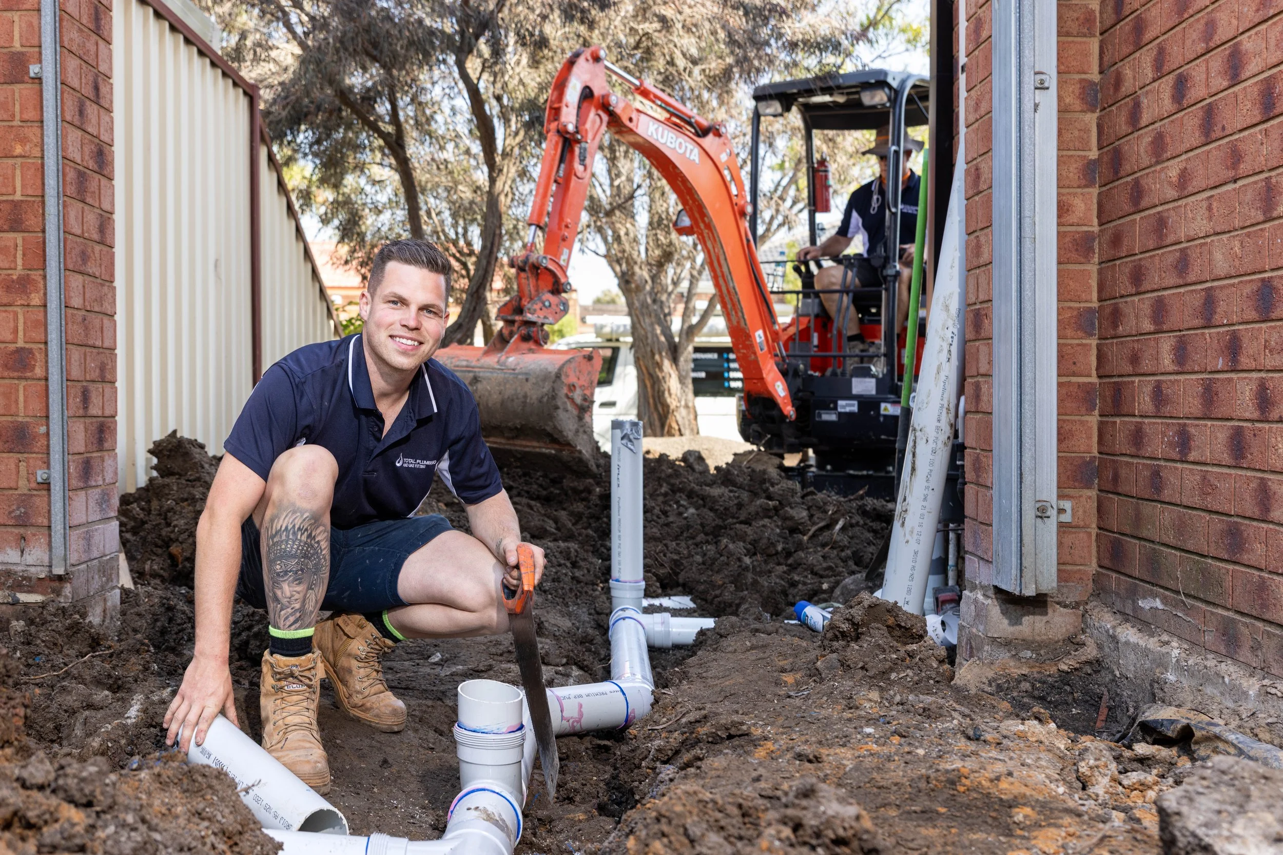 Two men working on installing plumbing pipes outside between brick walls, one kneeling and smiling at the camera, the other operating a small excavator in the background.