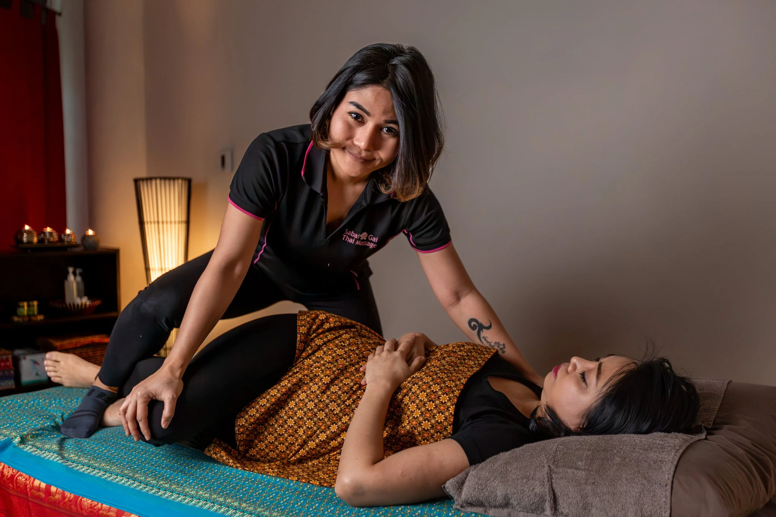 A woman giving a Thai massage to another woman lying on a mat in a spa room decorated with candles and warm lighting.
