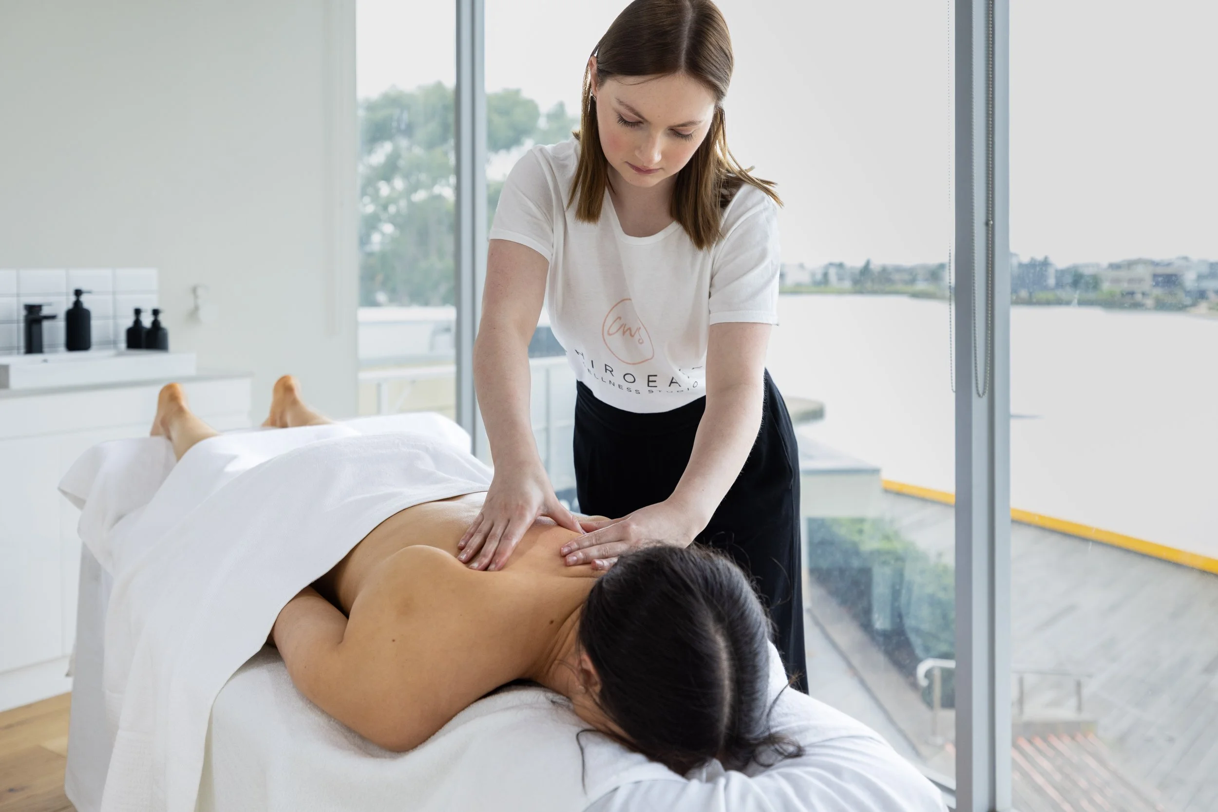 A woman receiving a massage from a professional therapist in a bright, modern massage room with large windows.
