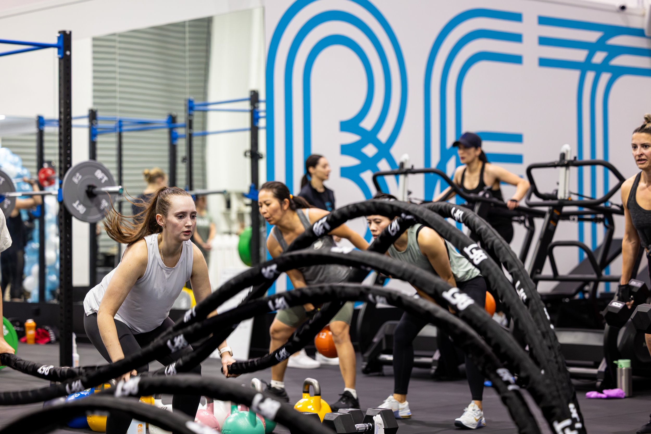 Women working out at a gym using battle ropes, with weight plates and kettlebells nearby, and large blue wall art in the background.