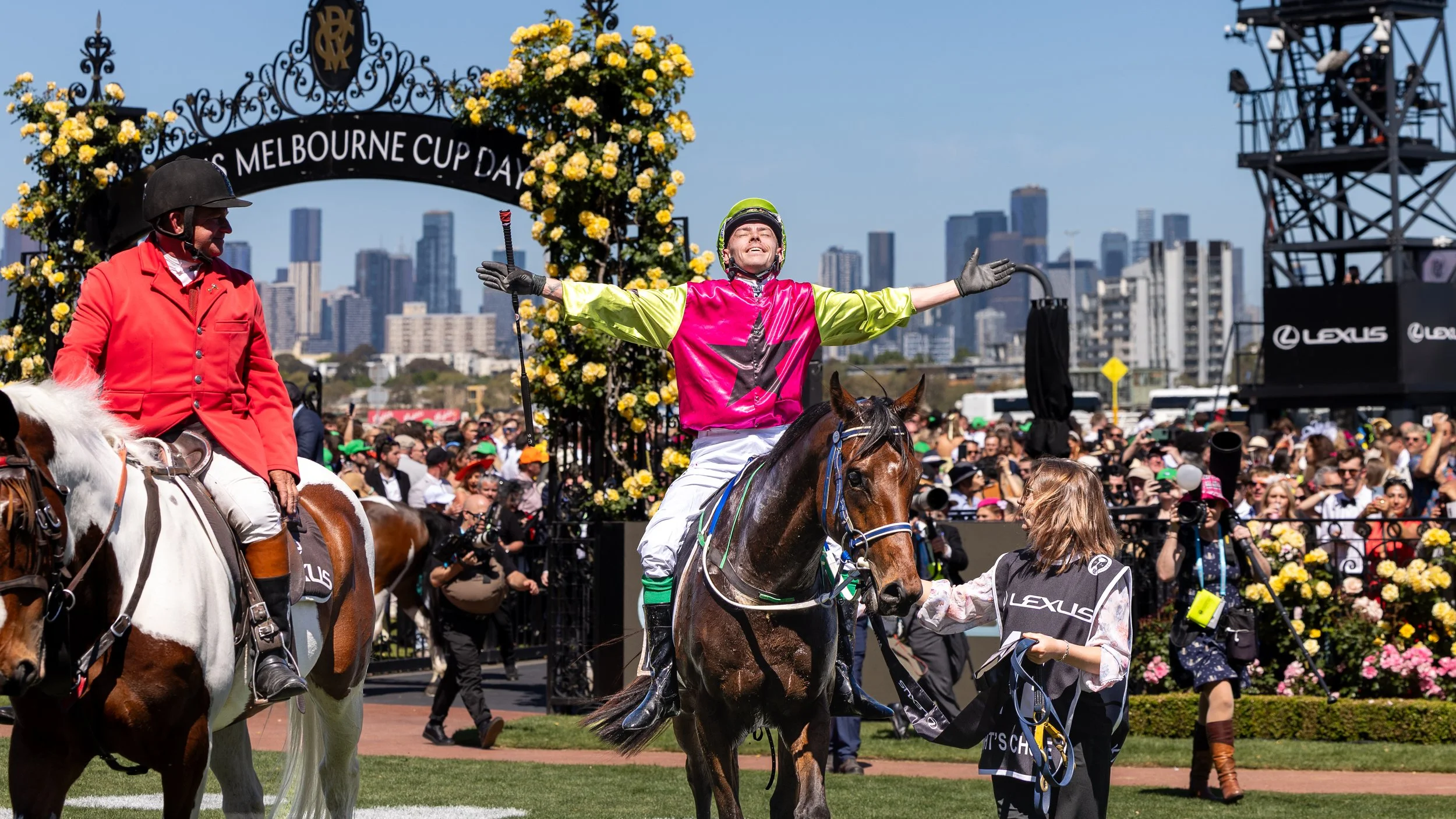 Racehorse jockey celebrating victory with arms outstretched on horseback at the Melbourne Cup Day event, surrounded by crowd, floral decorations, and city skyline in the background.