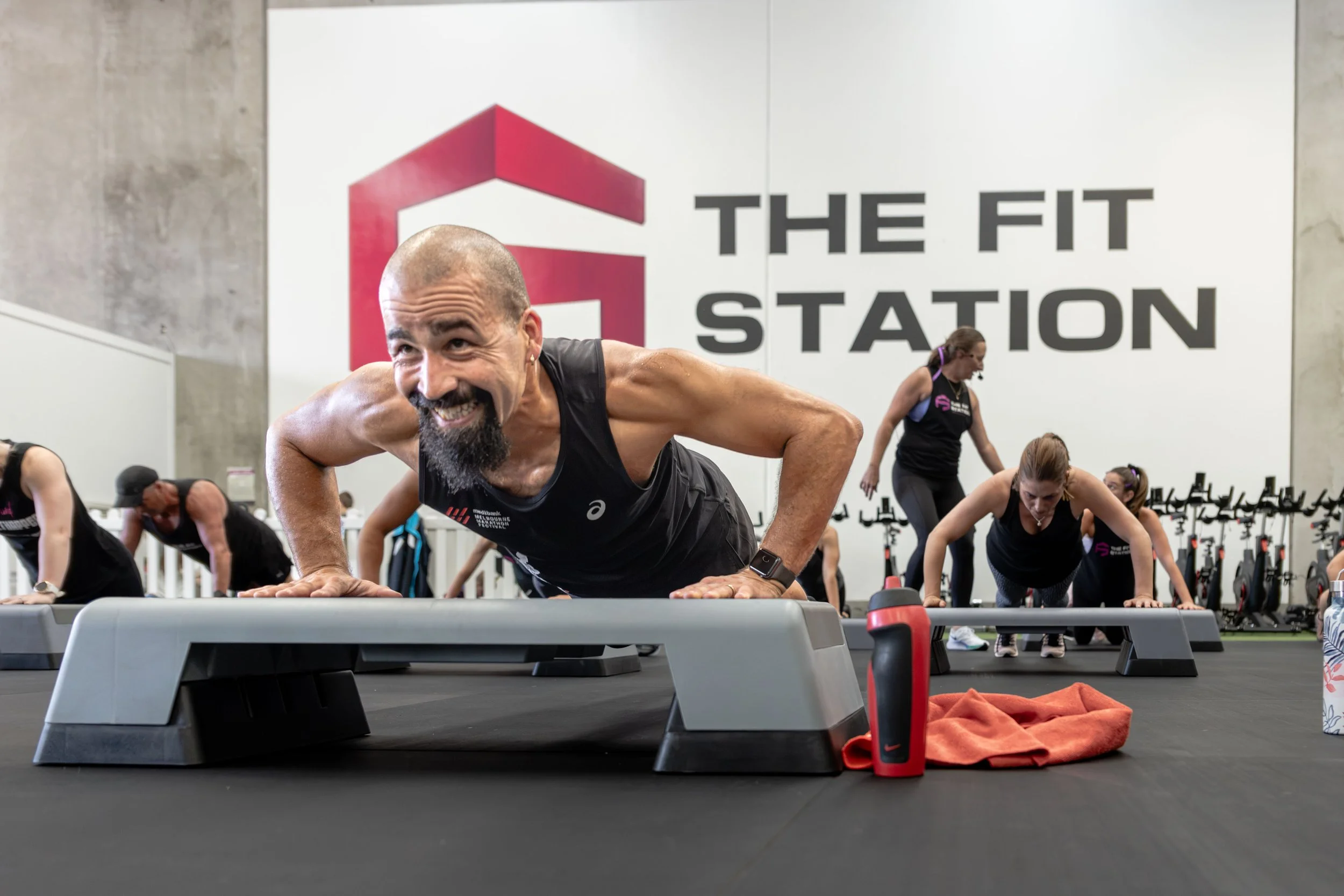 A group of people participating in a fitness class in a gym, with a man in the foreground doing push-ups on a step platform, smiling and wearing a black tank top. Other participants are in the background performing similar exercises, with a woman wea