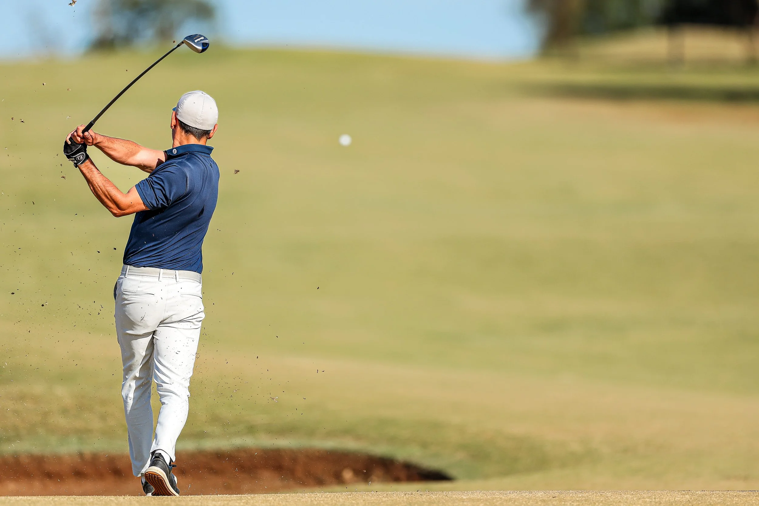 A golfer in a blue polo shirt, white pants, and a white cap swings a golf club on a golf course.