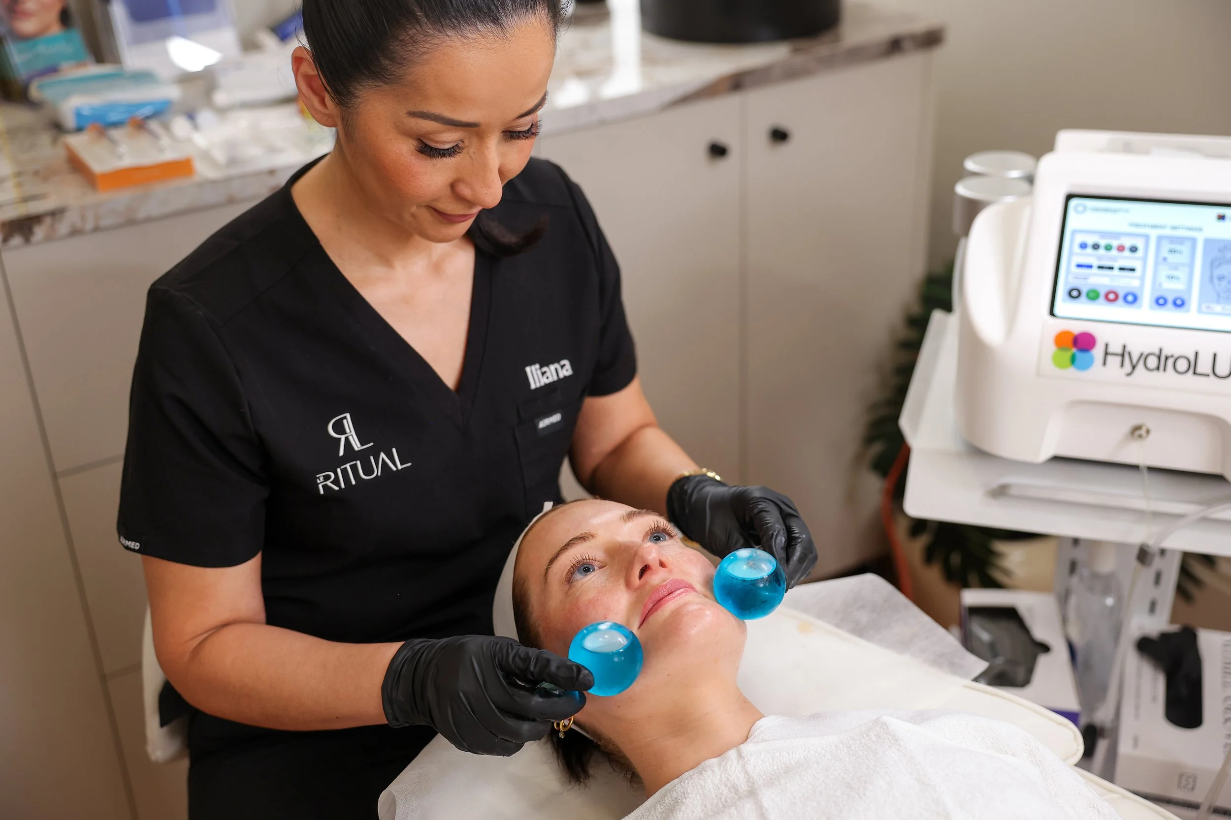 A woman lying on a treatment bed with a hydrodermabrasion device on her face, while a technician wearing black gloves and a black uniform with the name tag 'Iliana' performs a facial treatment. The technician is standing and looking down gently durin