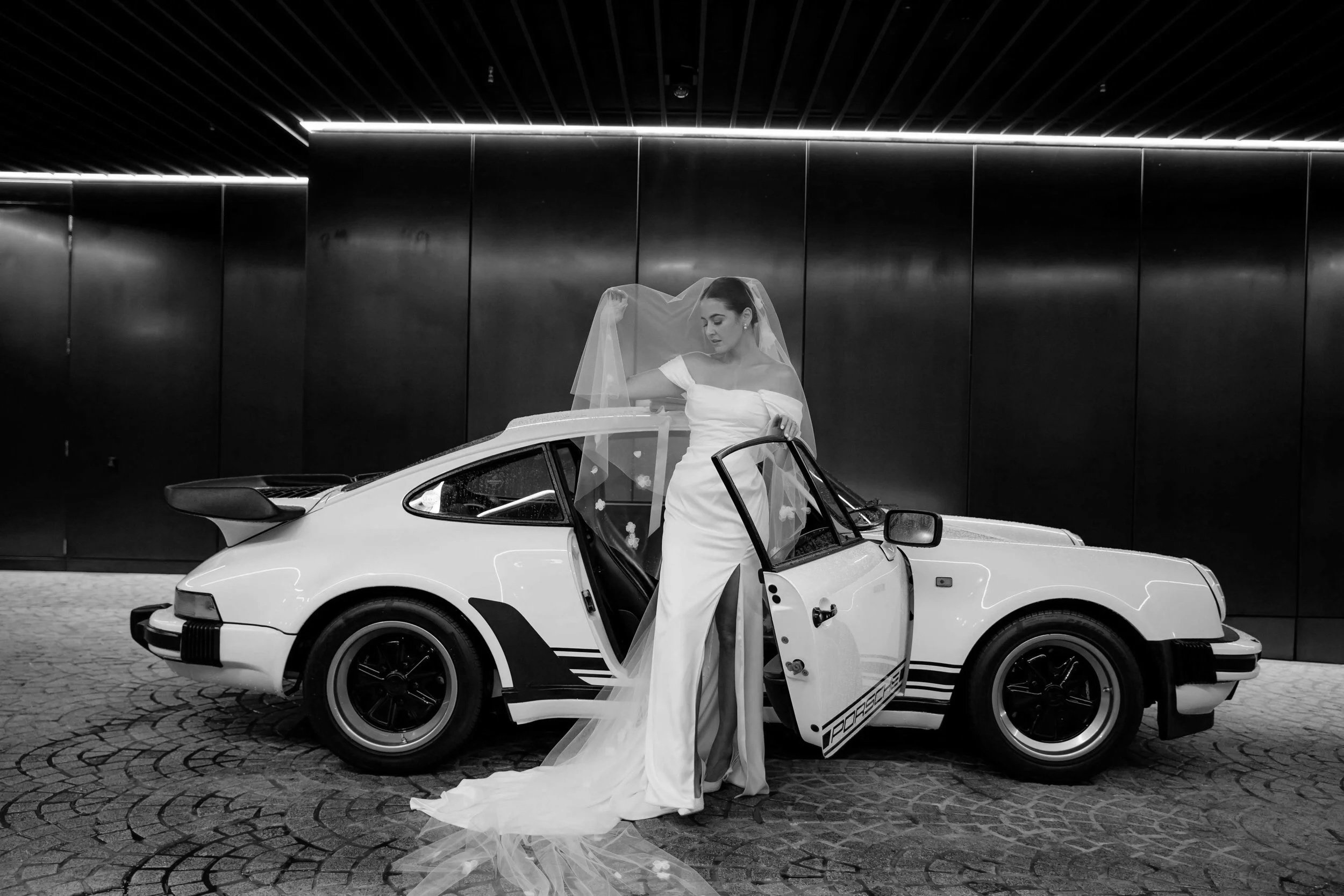 A bride in a wedding gown and veil standing next to a classic white Porsche sports car with open door, in an indoor setting with dark walls and shining ceiling.