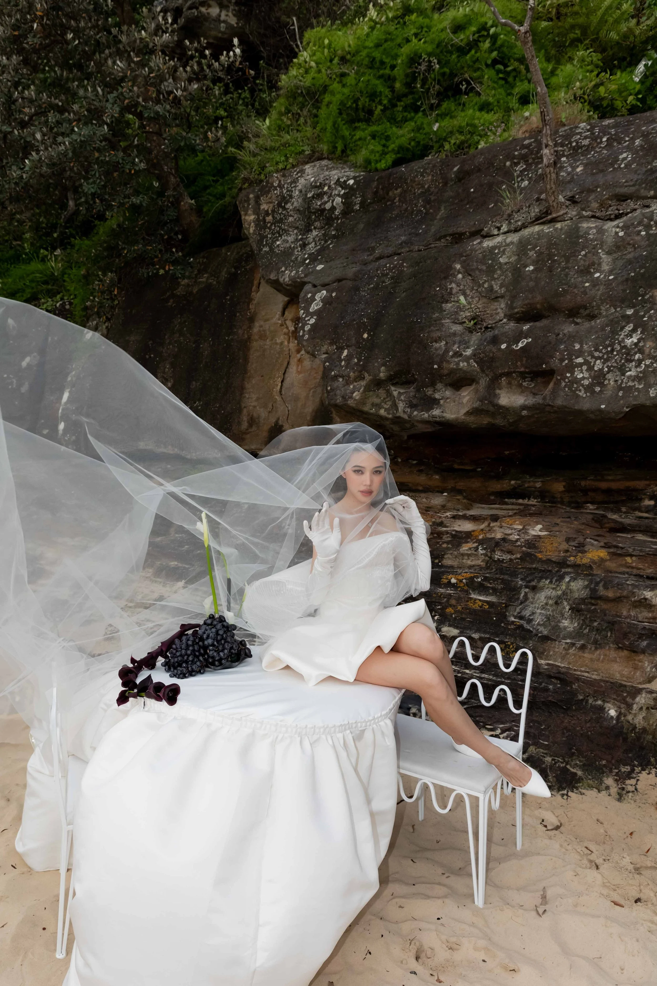 A woman in a white dress and gloves sitting on a white table with a veil over her face, holding a bunch of grapes, on a sandy surface with rocks and greenery in the background.