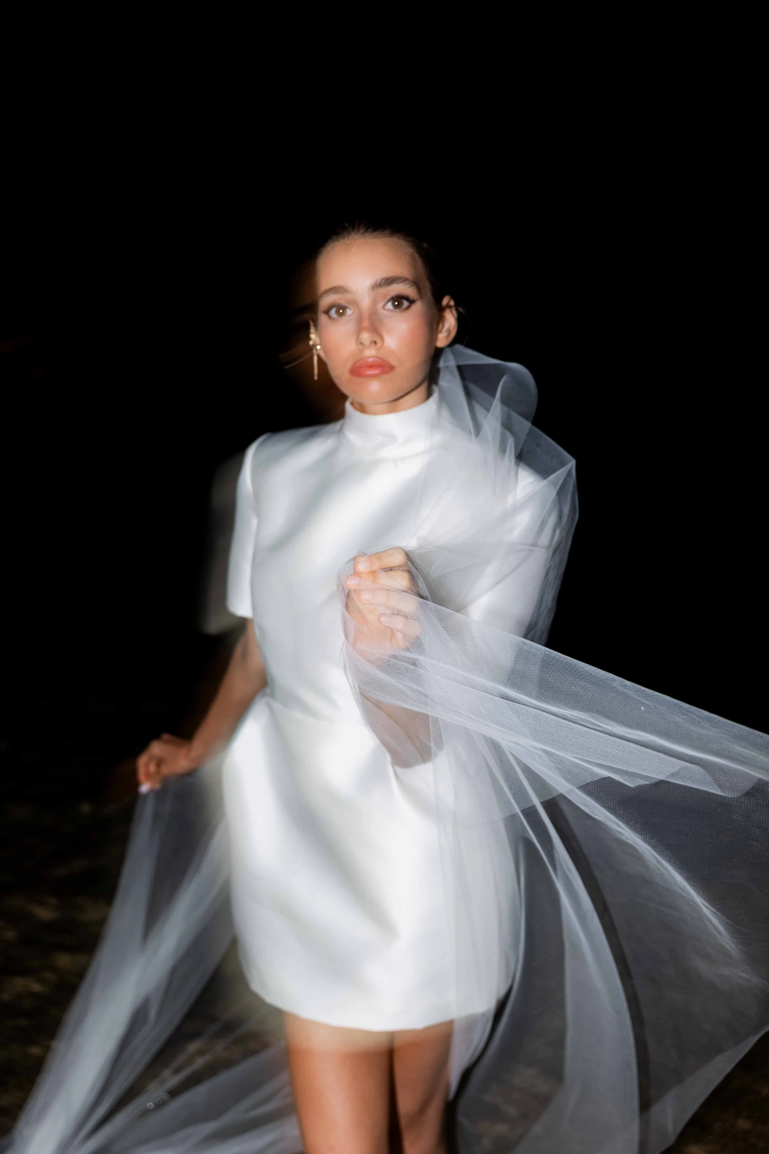 Woman in a white dress holding a sheer veil, standing against a black background.