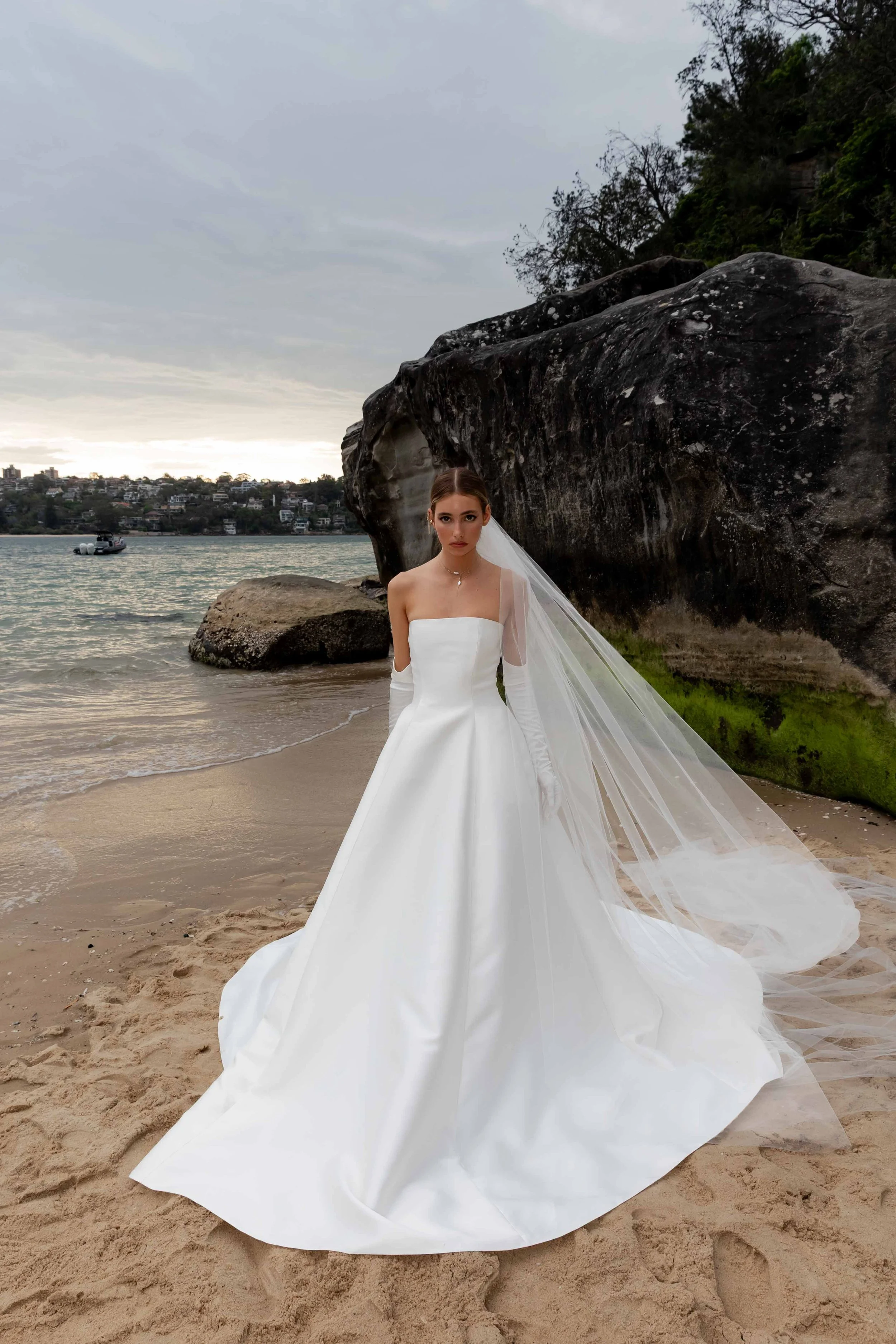 A bride in a white wedding gown and long veil standing on a beach near large rocks and water, with a cloudy sky overhead.