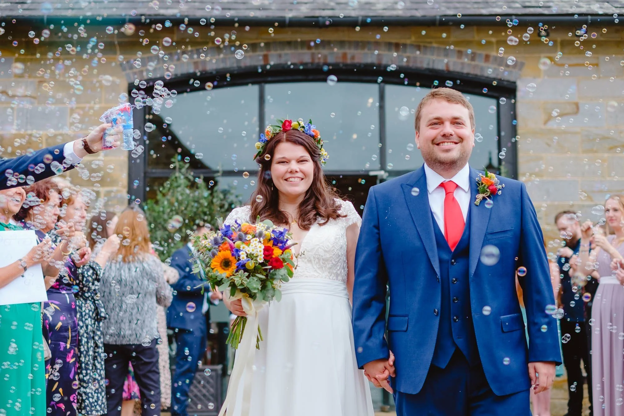 A newlywed couple, holding hands outside Hendal Manor Barns, Uckfield, smiling as guests celebrate with bubbles.
