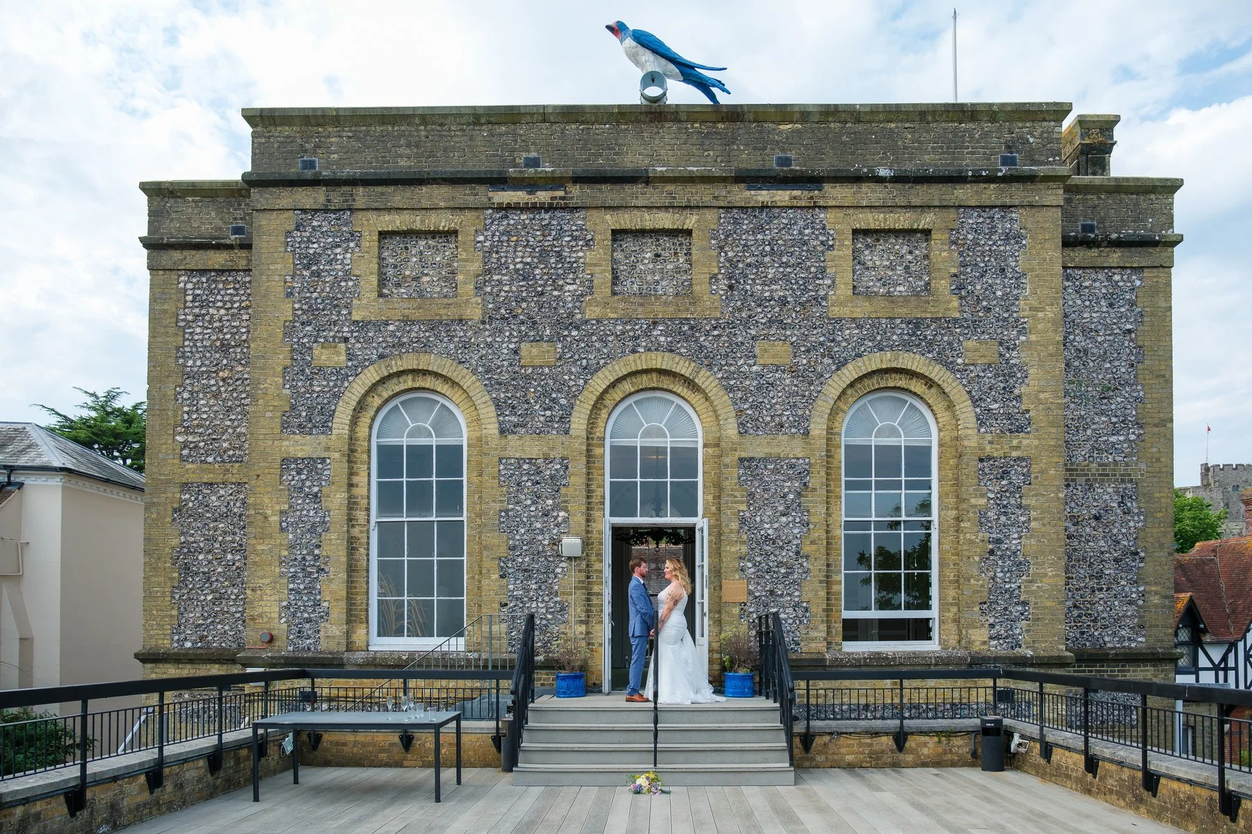 A bride and groom standing face to face on the steps of Arundle Registry Office with large arched windows, holding hands, with a bouquet on the ground in front of them, under a partly cloudy sky.