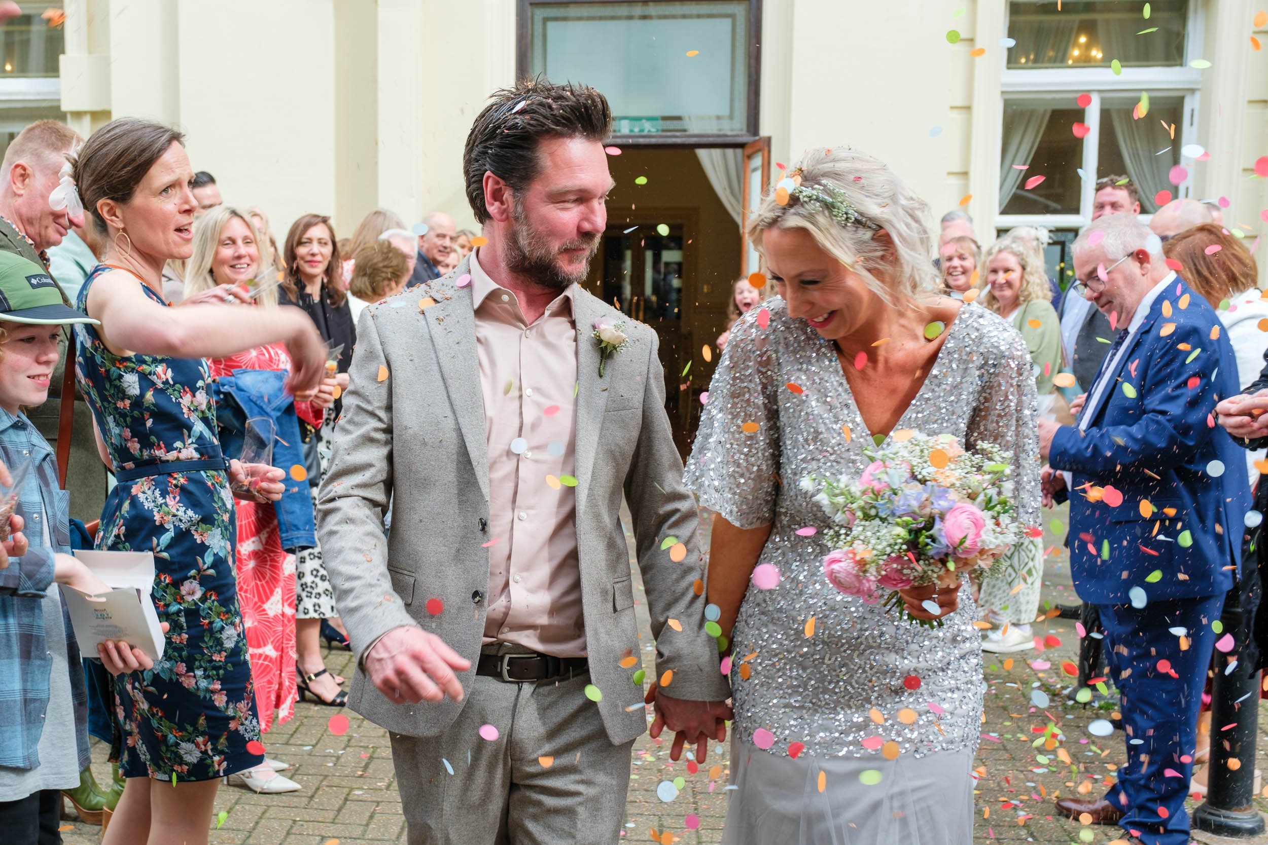 Couple holding hands and smiling surrounded by guests at a wedding celebration with falling confetti.