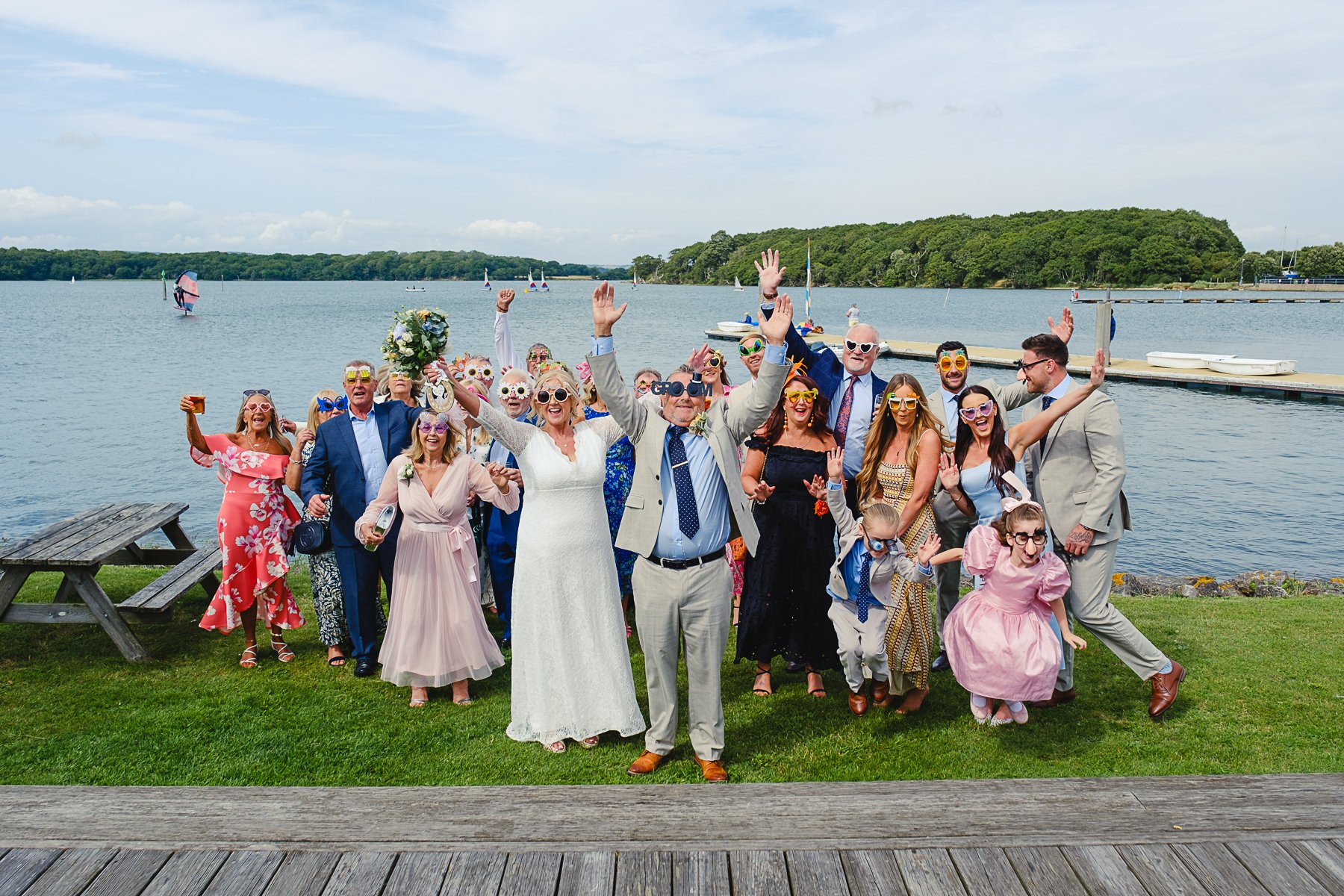 A group of people in colorful outfits celebrating by a lake, wearing quirky sunglasses, with some raising their hands, and a few kids in the front, on a grassy area near a wooden deck, with boats and sailboats in the water and a green tree-lined shor