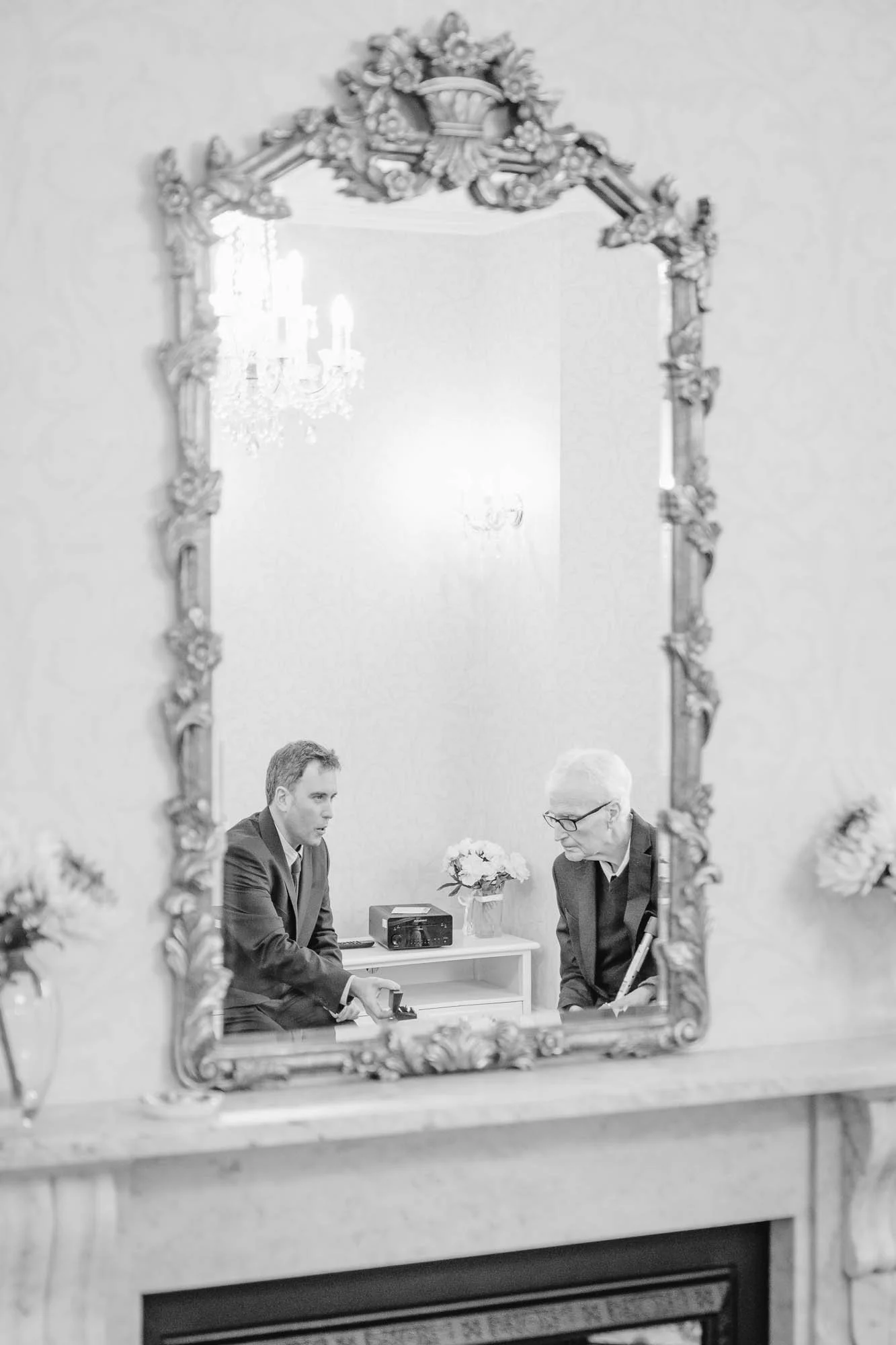 A black-and-white photo shown through an ornate mirror frame in a ceremony room at The mansion House Tunbridge Wells Registry Office, reflecting two men and the groom is showing his Dad the wedding rings.