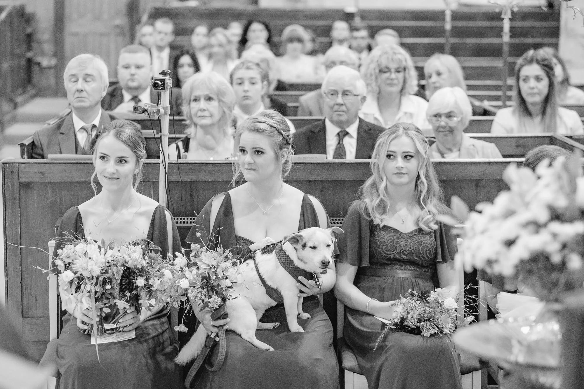 Black and white photo of three young women in formal dresses sitting in the front row of Rotherfield St Denys Church. One of the women has a dog on her lap. There are flowers in front of them and a congregation of people sitting in the pews behind th