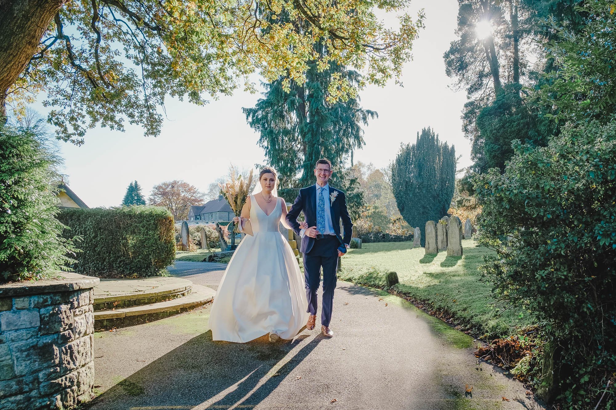 Bride and groom walking down the path to All Saints Church Crowborough on a sunny day, surrounded by trees and gravestones.